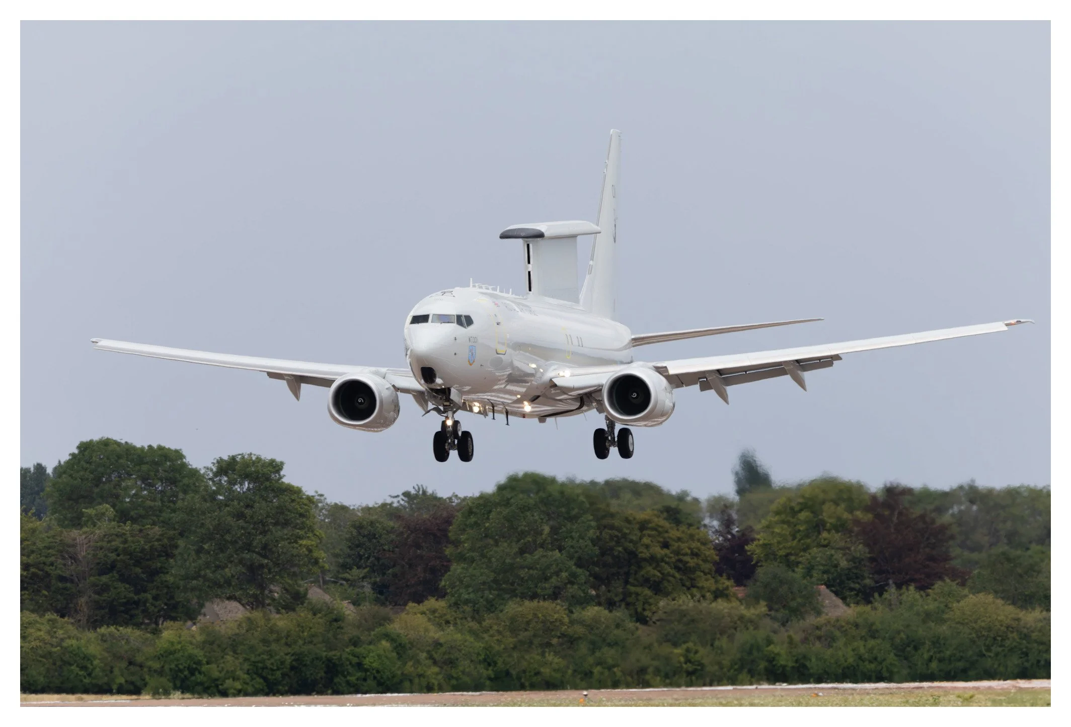 An aircraft in flight, descending with its landing gear extended, above a landscape of trees and an overcast sky.