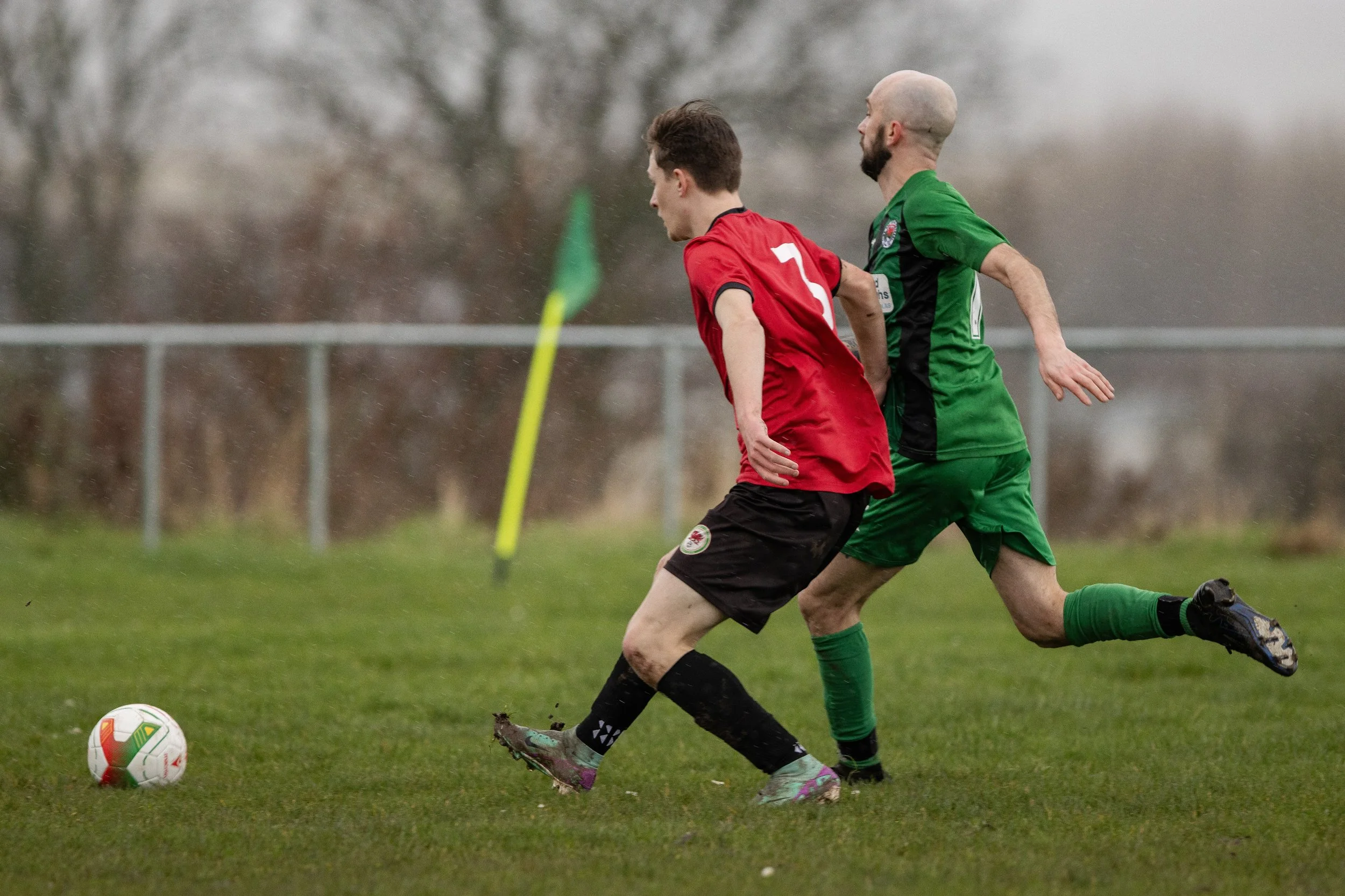 Two soccer players competing for the ball on a wet field, one in red jersey and black shorts, the other in green jersey and shorts, with trees and a fence in the background.