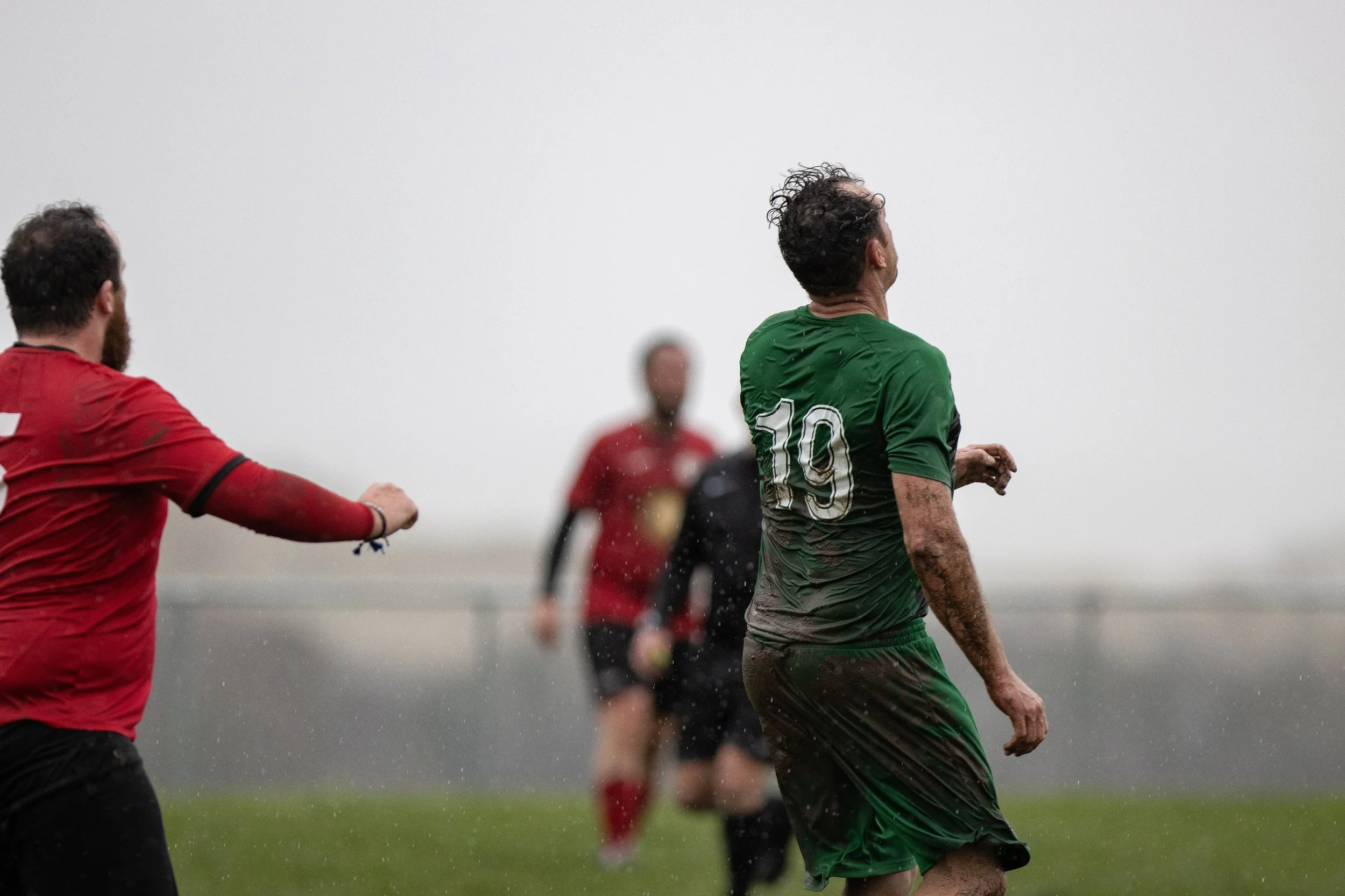 A soccer player wearing a green jersey with the number 19 on the back, covered in mud, is in action on a rainy field. Two other players in red jerseys are nearby, one with his arm extended, and a blurred referee or player in the background.