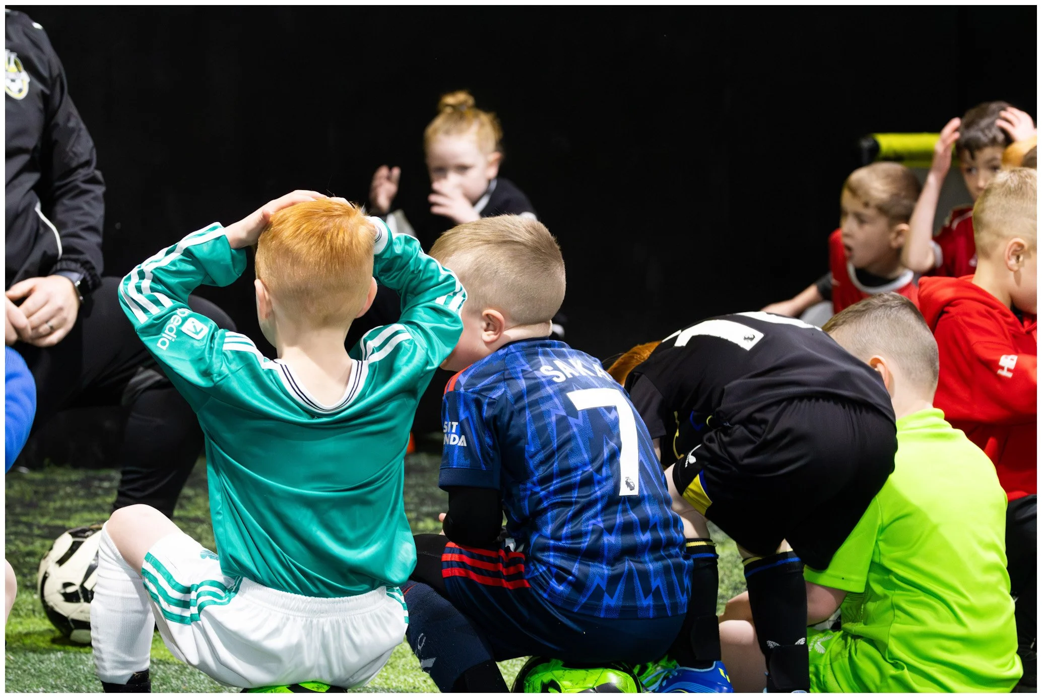 Young soccer players sitting on the field during a team meeting, with a coach standing nearby, wearing casual sportswear