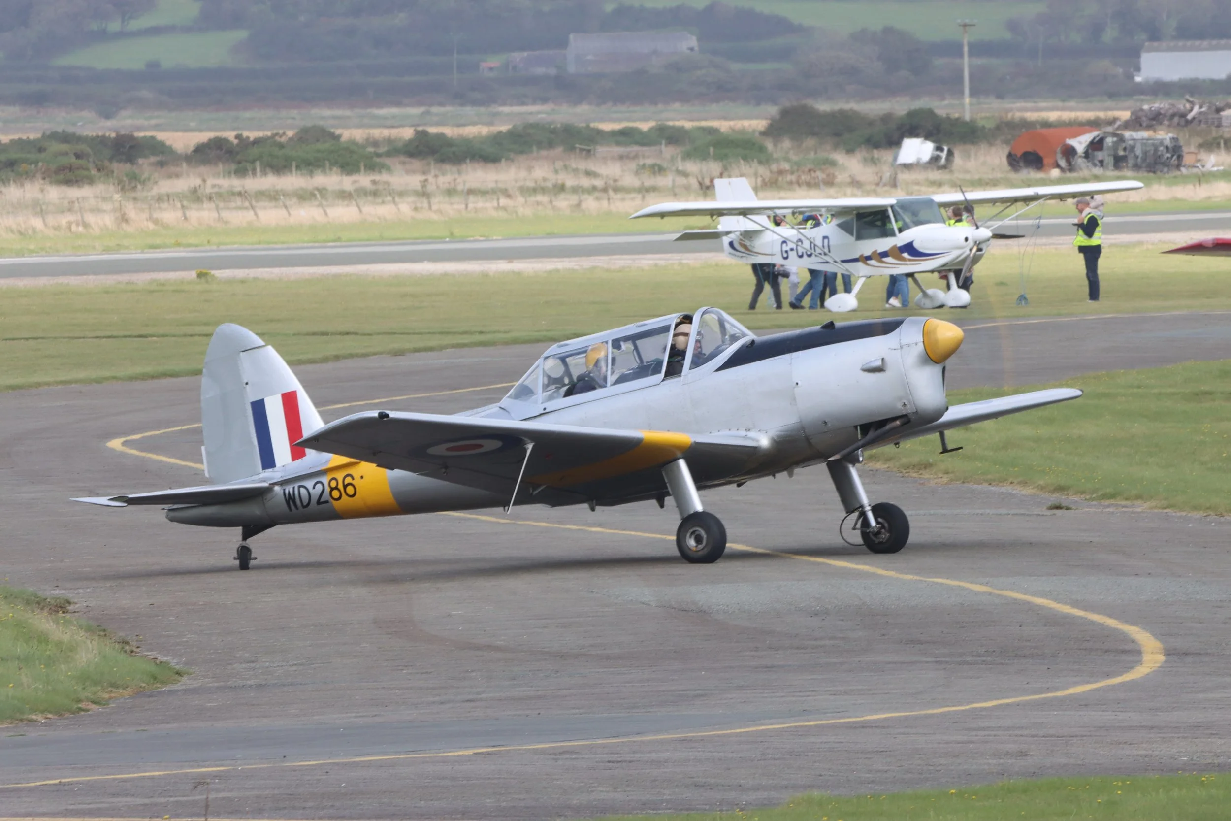 A vintage aircraft with a silver body, black and yellow accents, and a red, white, and blue roundel on the side, taxis on a runway. In the background, a small white and blue plane with a tail number G-CGUJ is surrounded by people, and two rusty, wrec