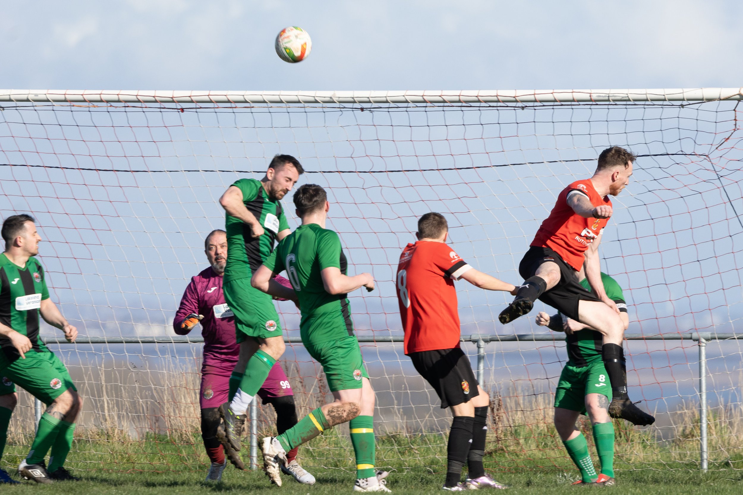 Soccer players in green and red jerseys jumping and attempting to head the ball during a match on a grassy field with a goal net in the background.