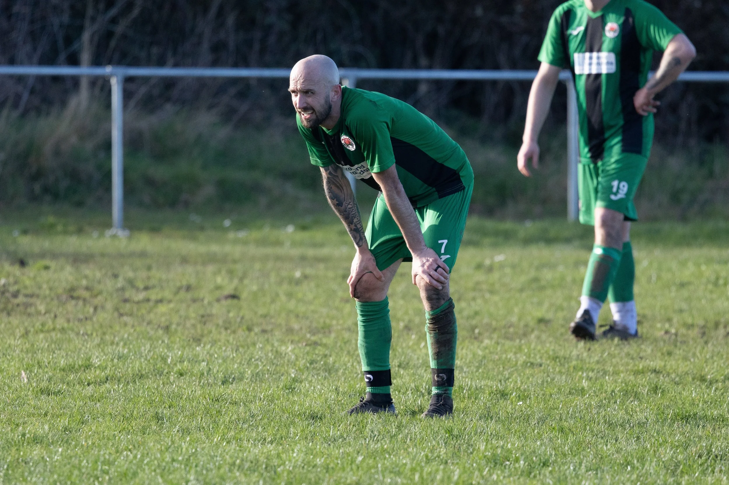 A soccer player dressed in a green jersey with the number 7, bent over holding his knee during a game on a grassy field.