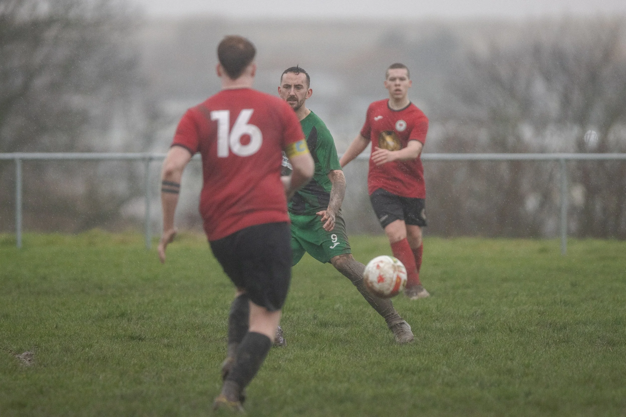 Three soccer players on a grassy field in a rainy weather. One in a green jersey with the number 9 appears to be kicking the ball, while two players in red jerseys with the numbers 16 and 8 are nearby, watching the play.