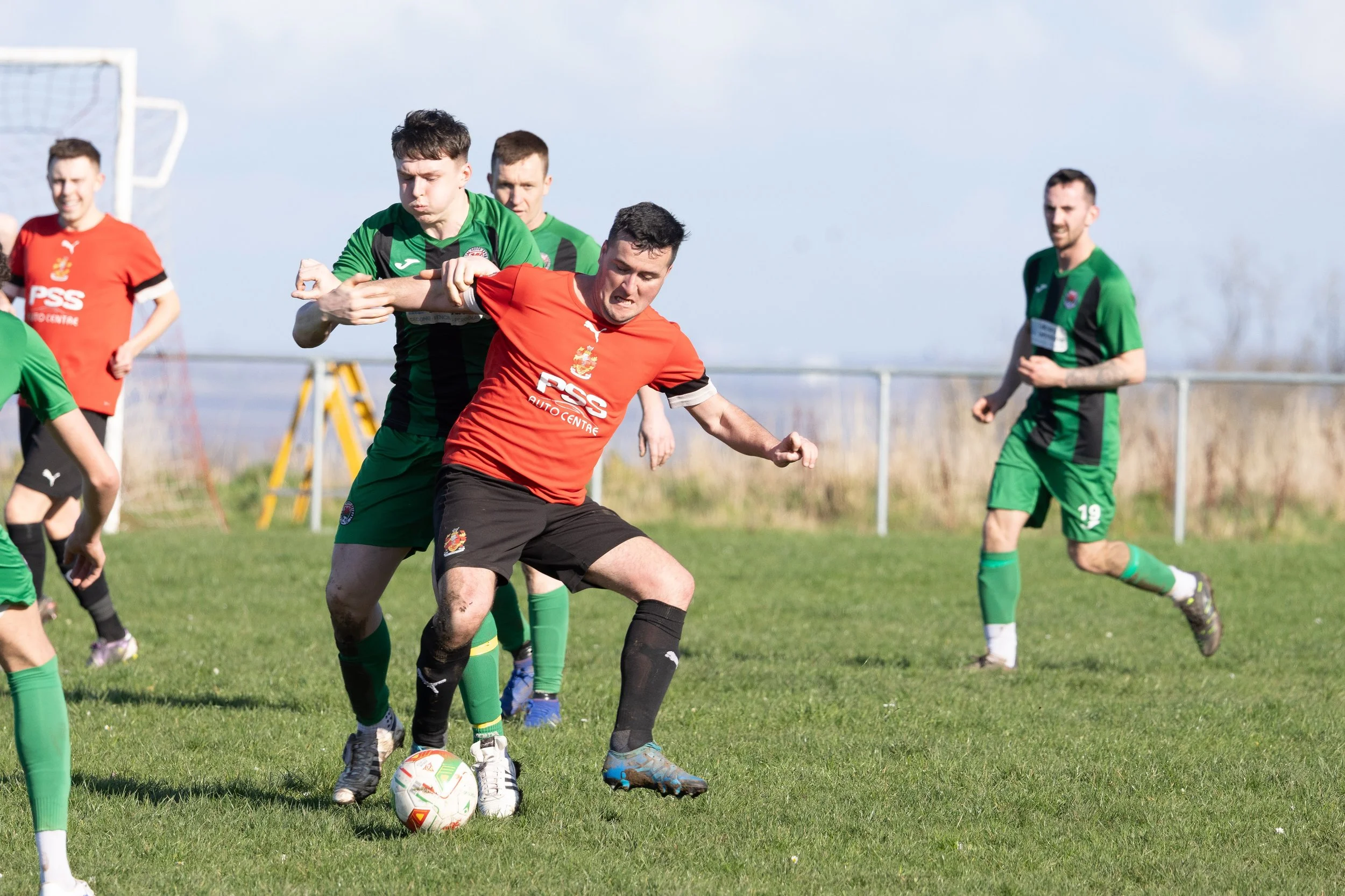 Soccer players competing for ball on a grassy field with a goalpost in the background.