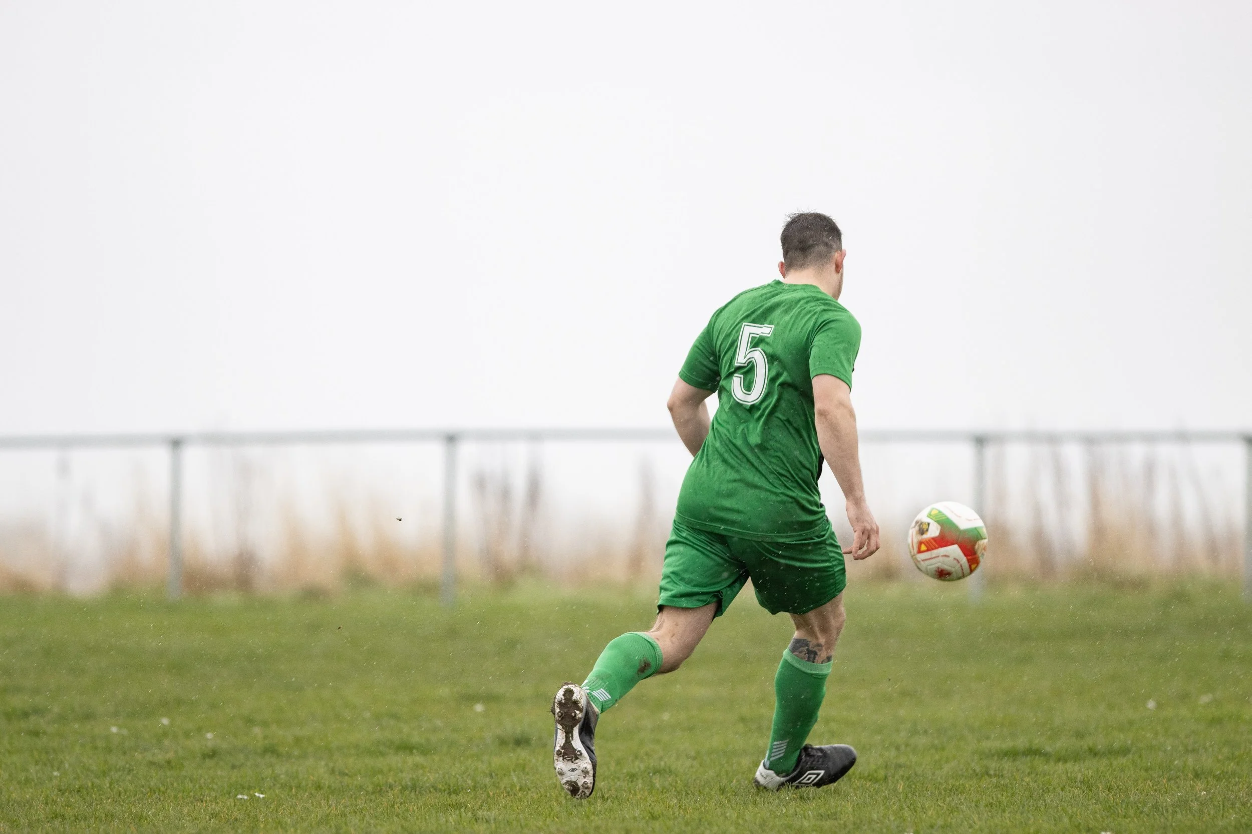 A soccer player in a green uniform with the number 5 on his back, kicking a soccer ball on a grassy field with a blurred background and overcast sky.