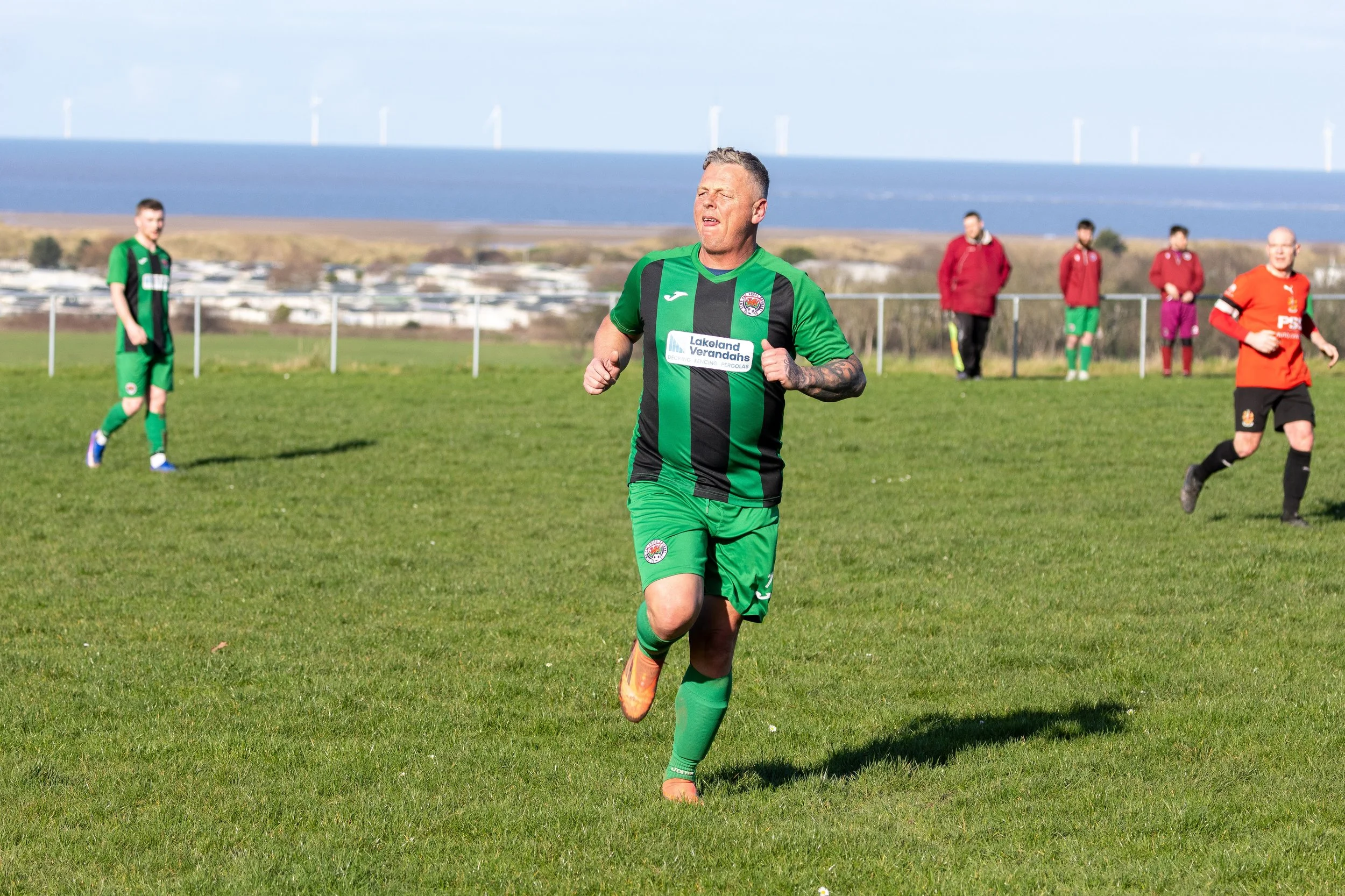 A man in a green and black soccer uniform runs on a grassy field during a match. Other players are visible in the background, with a scenic landscape and wind turbines in the distance.