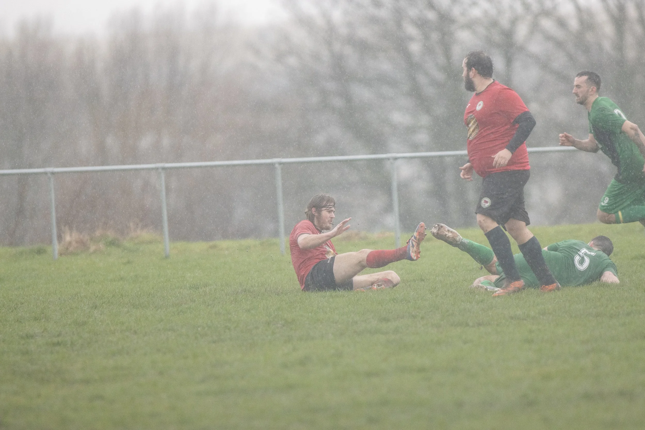 Rainy soccer match with players in red and green uniforms on the field, some players on the ground, rain falling, and background of trees and fence.