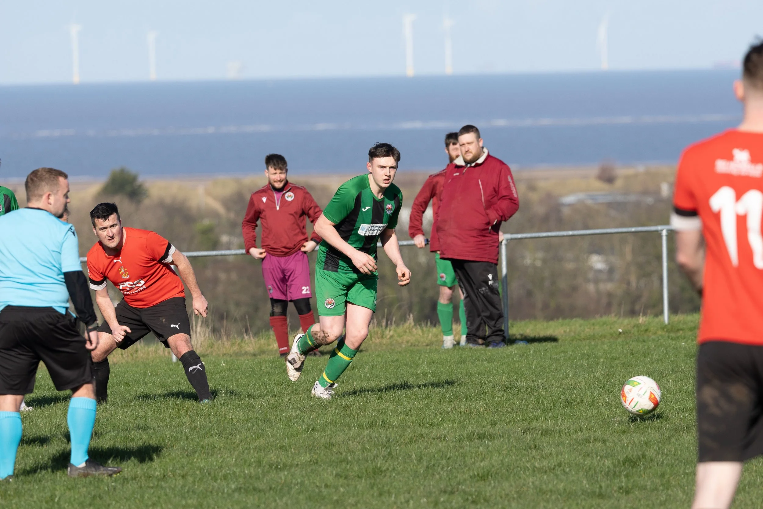 Soccer players on a green field during a match, with a ball in the foreground and a backdrop of wind turbines and a blue sky.