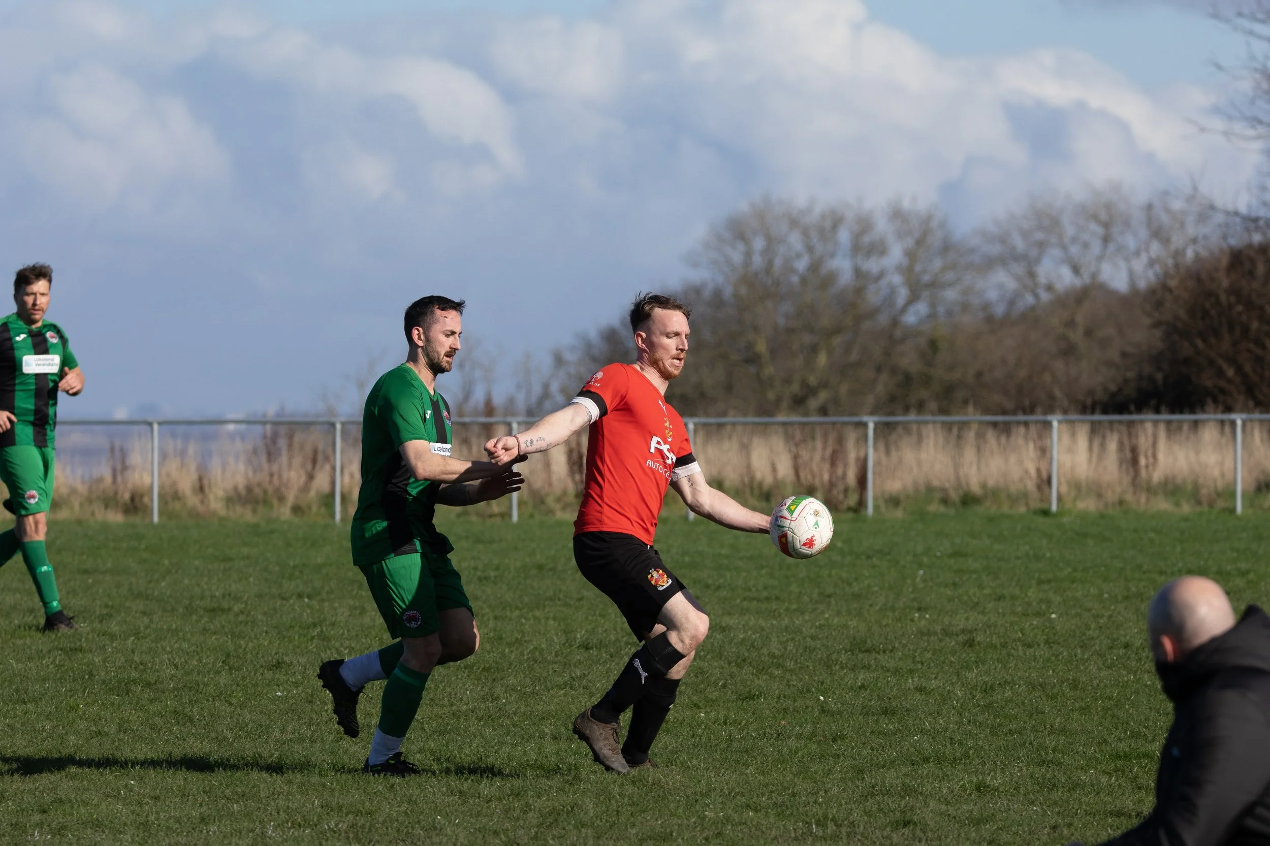 Soccer players on a field, one in a red jersey with black shorts controlling the ball, while others in green jerseys are around him during a match.
