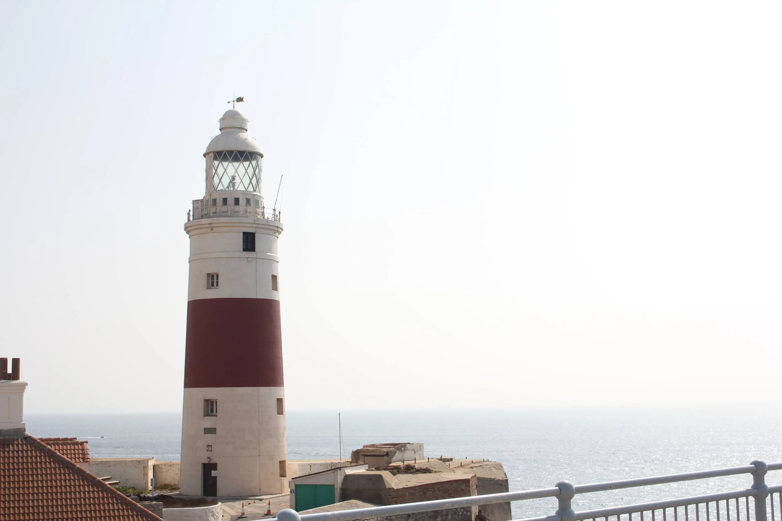 A tall lighthouse with a red band around its middle, situated near the ocean with a clear sky and calm sea in the background.