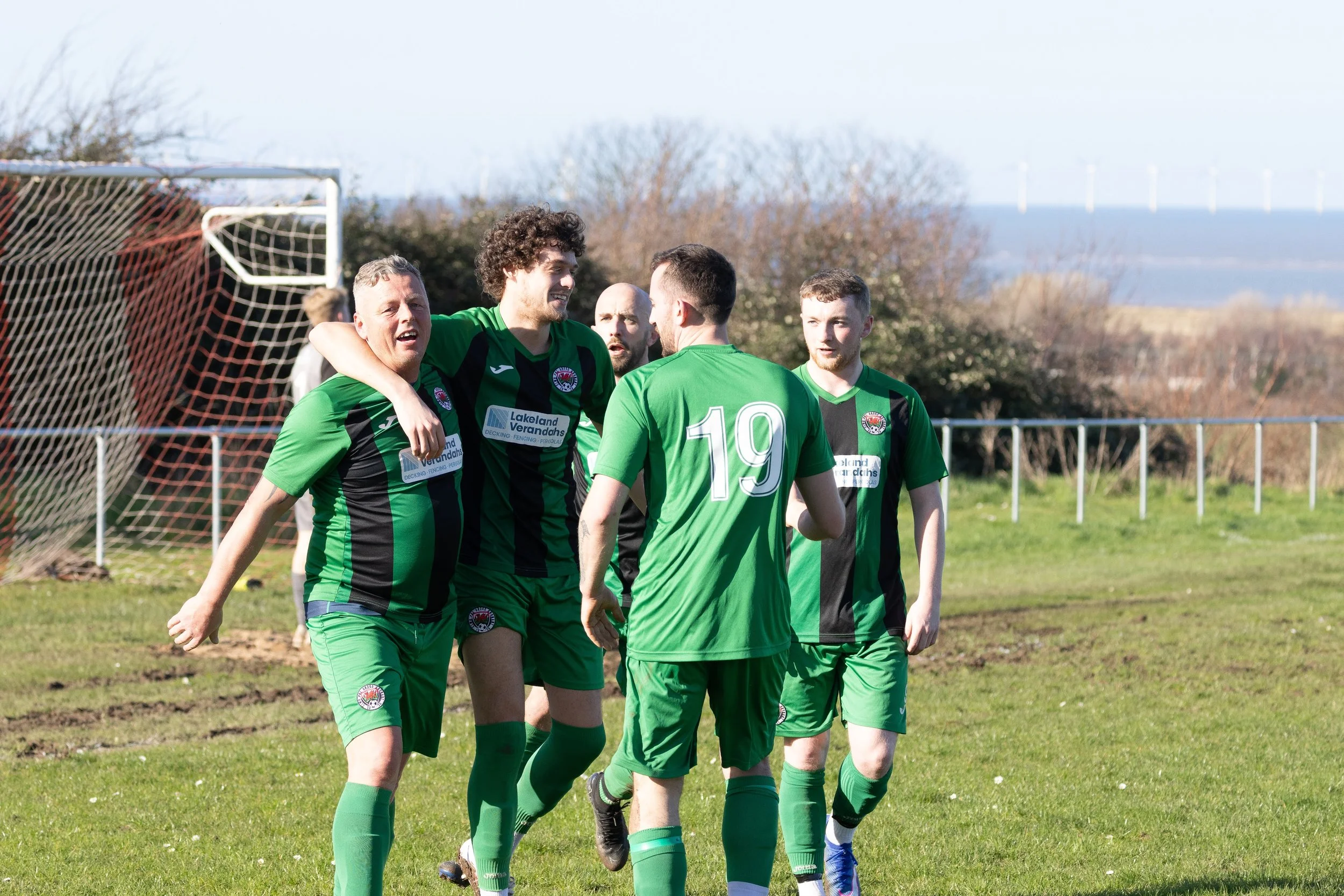 Soccer players in green uniforms celebrating after scoring a goal on a grassy field, with a goalpost and wind turbines visible in the background.