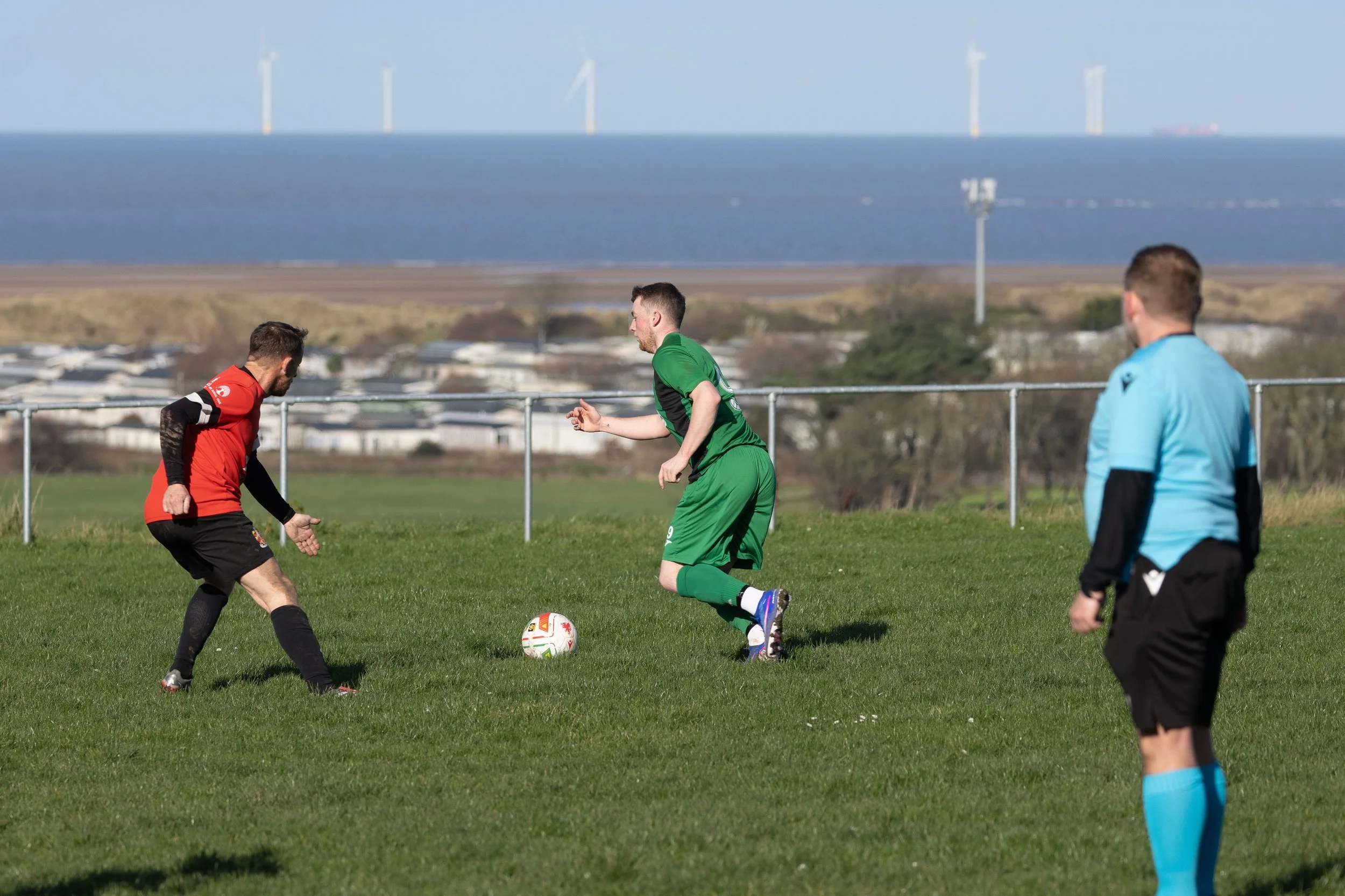 Soccer match in progress with three players and a referee on a grass field, with wind turbines and water in the background.