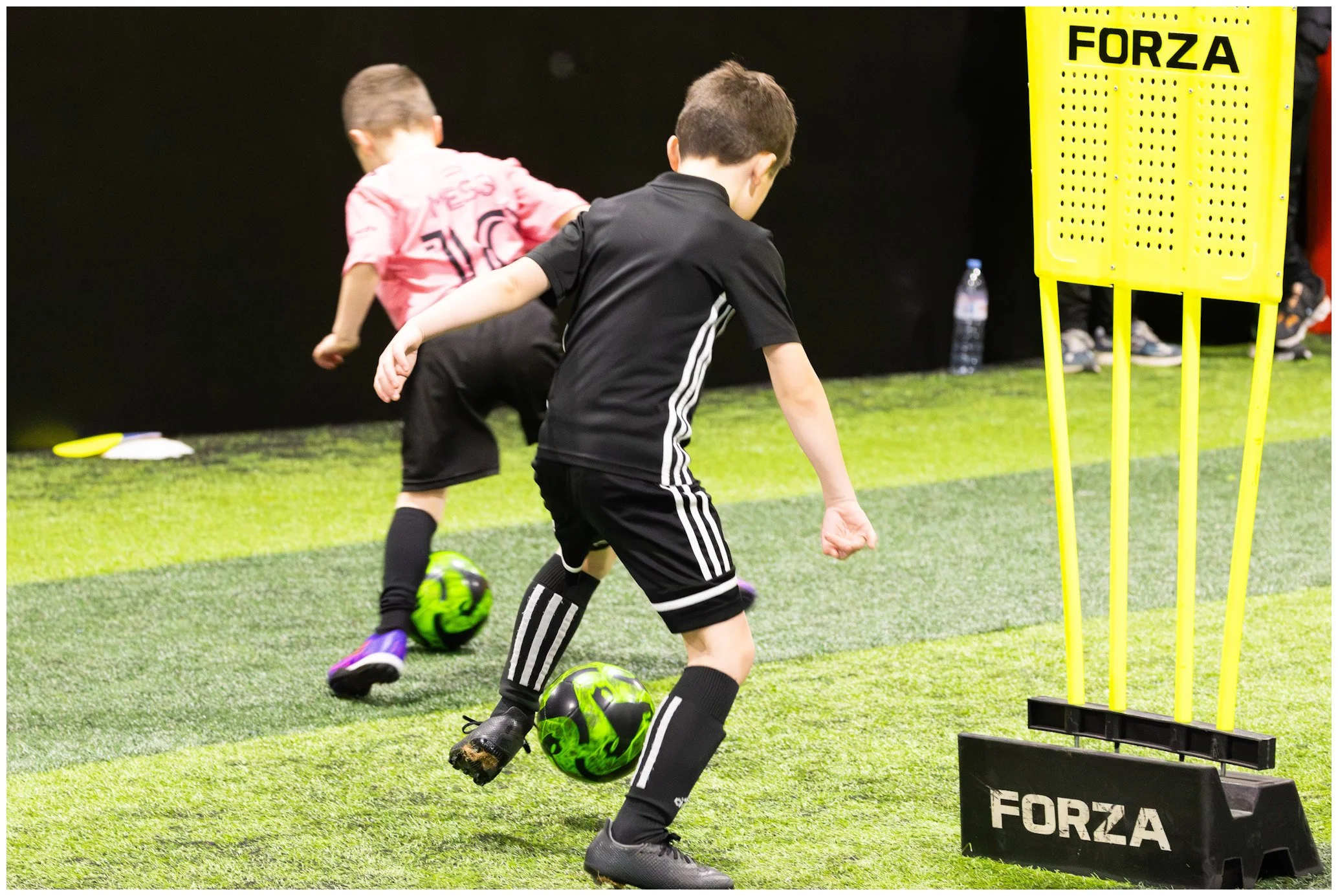 Two young boys playing indoor soccer, practicing dribbling skills with green soccer balls, on artificial turf with training equipment nearby, including a yellow training mannequin labeled 'FORZA'.