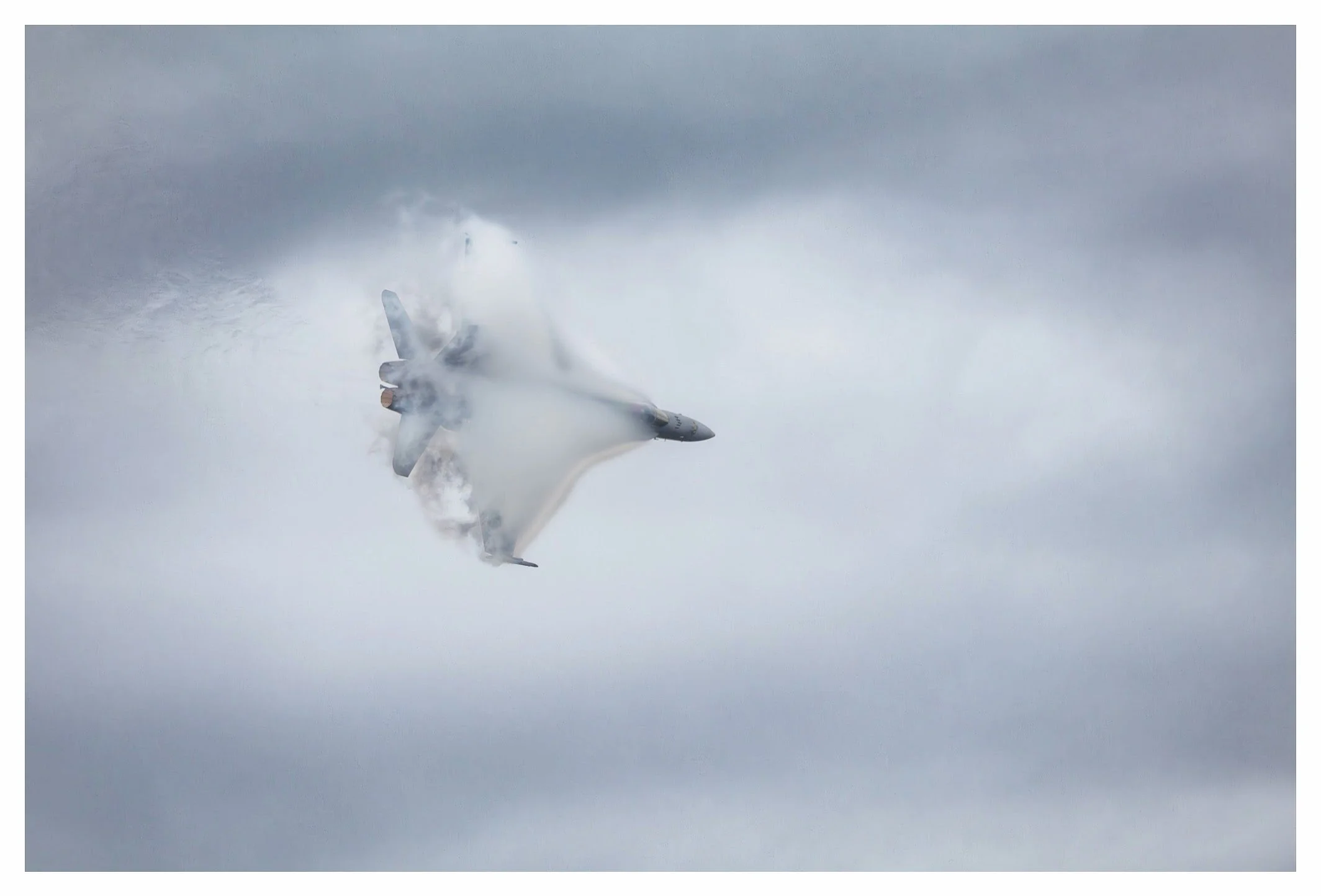 A military jet flying at an angle through cloudy skies, leaving a vapor trail behind.