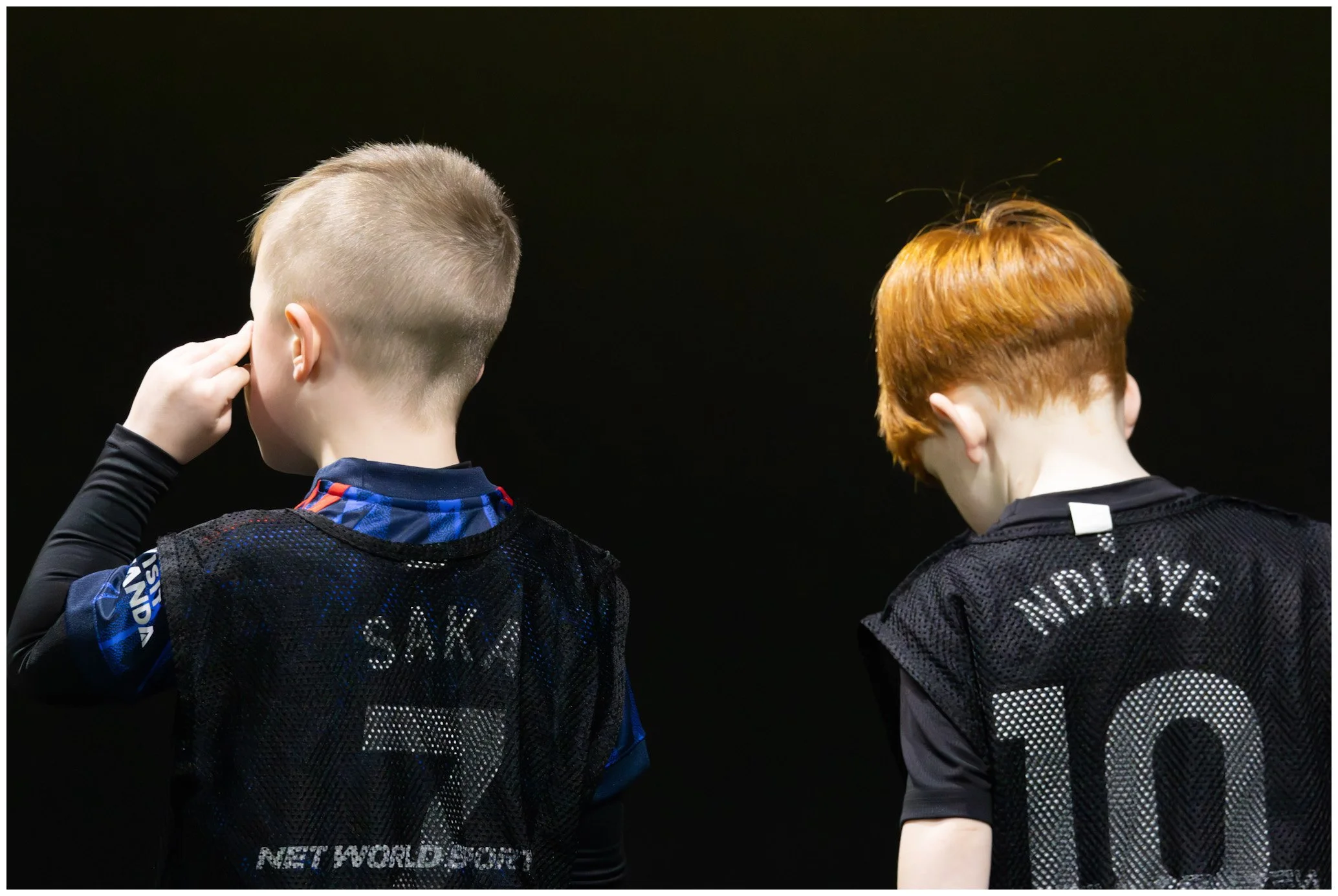 Two young boys wearing black jerseys with names and numbers on the back, standing with their backs to the camera, against a dark background.