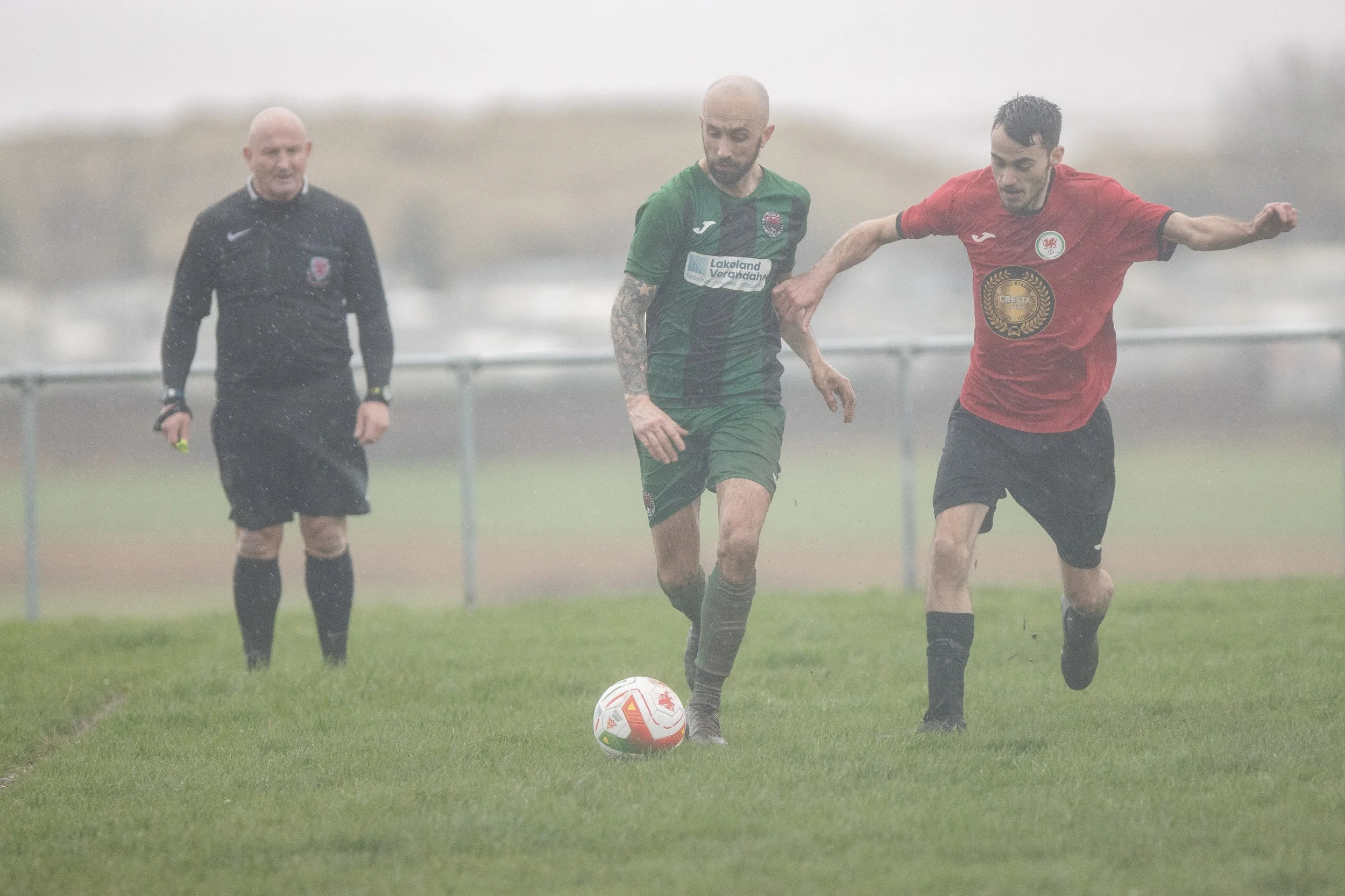 Two soccer players competing for the ball on a rainy field, with a referee in the background.