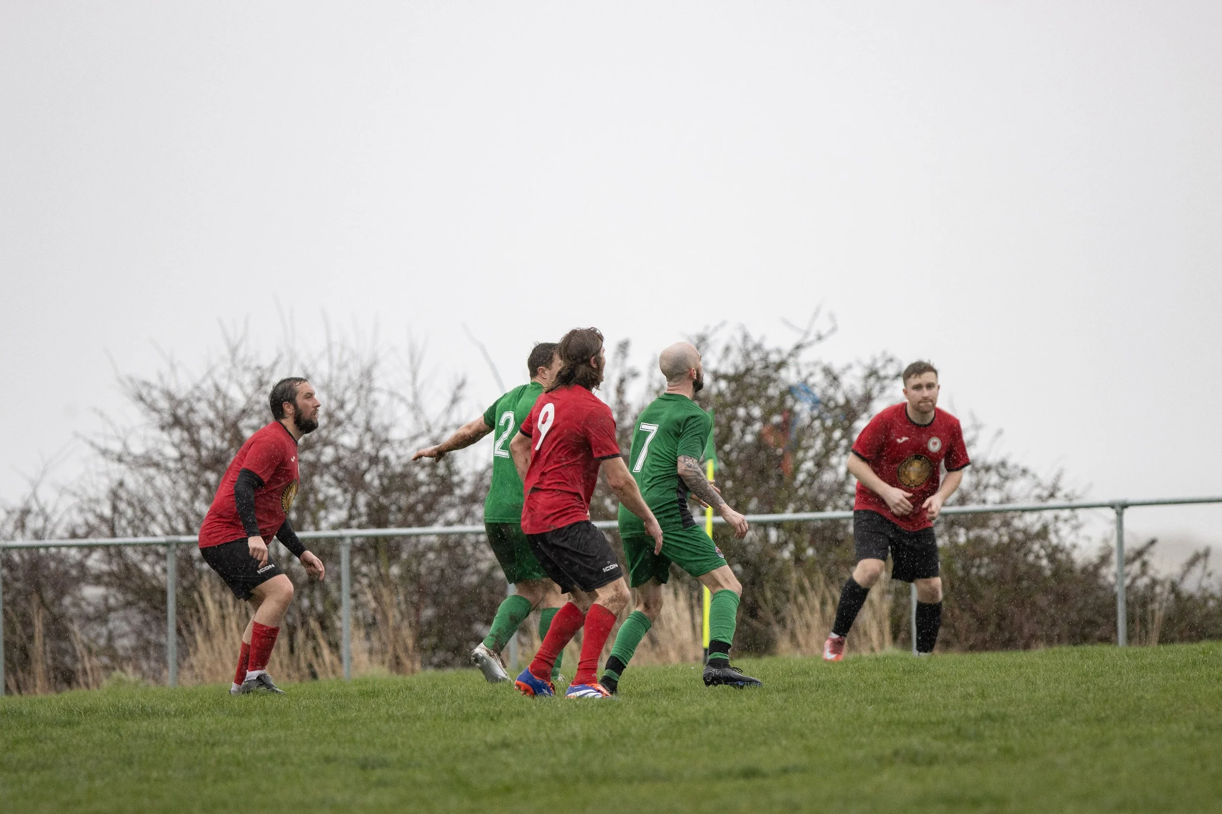 Soccer players in red and green jerseys playing on a grassy field on a cloudy day