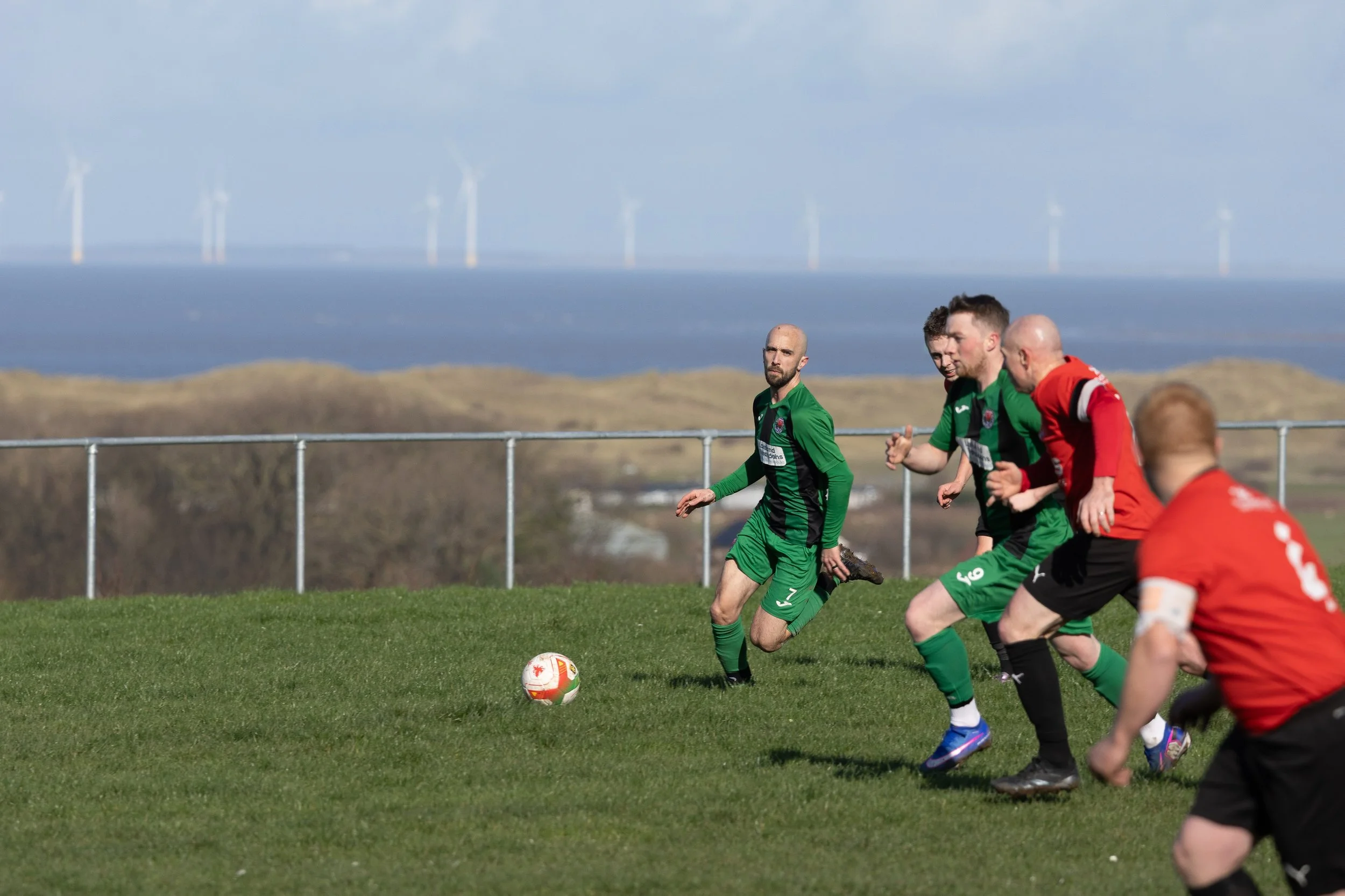 A group of soccer players in green and red jerseys running towards a soccer ball on a grass field with wind turbines on a hill in the background.