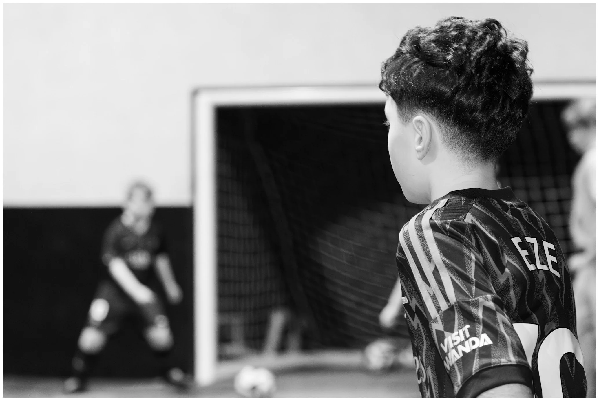 A young soccer player with curly hair looking at a goalie in front of a soccer goal, in black and white.