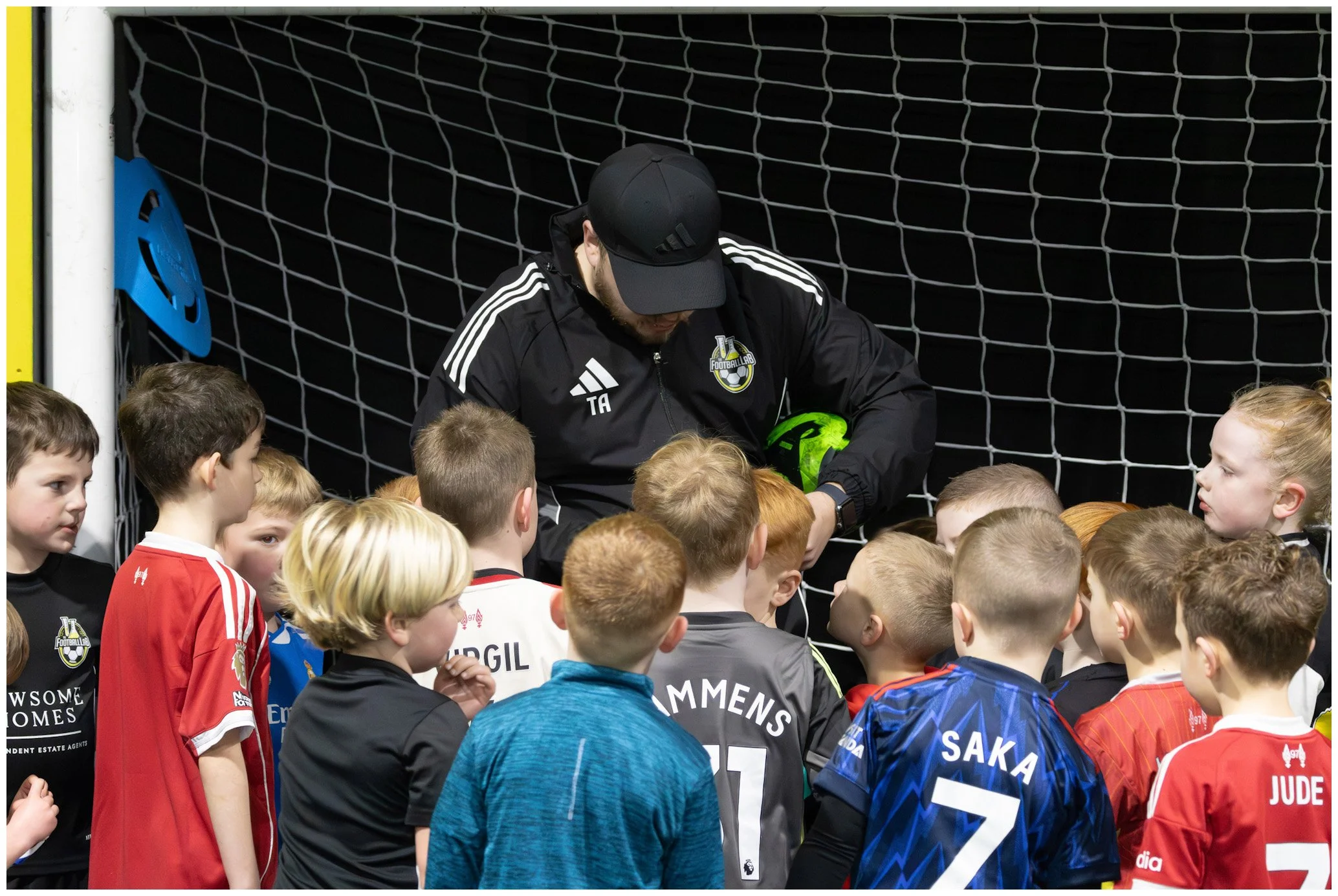 A group of children gathered around a soccer goalkeeper who is sitting on a bench behind a goal net, holding a green soccer ball and receiving instructions or a pep talk.