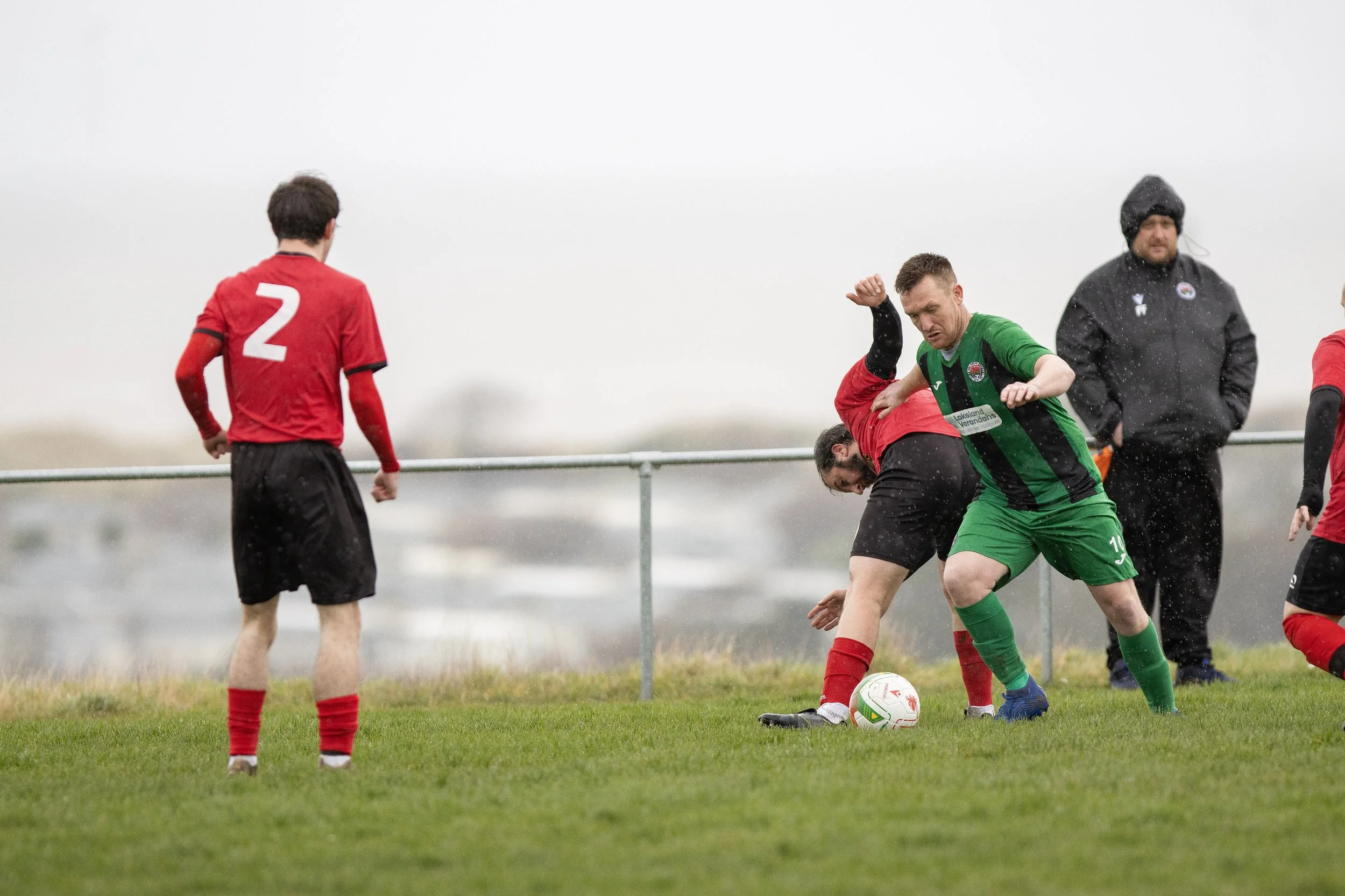 Two soccer players in red and green uniforms compete for the ball during a match on a grassy field in rainy weather, with a referee and wet conditions visible.
