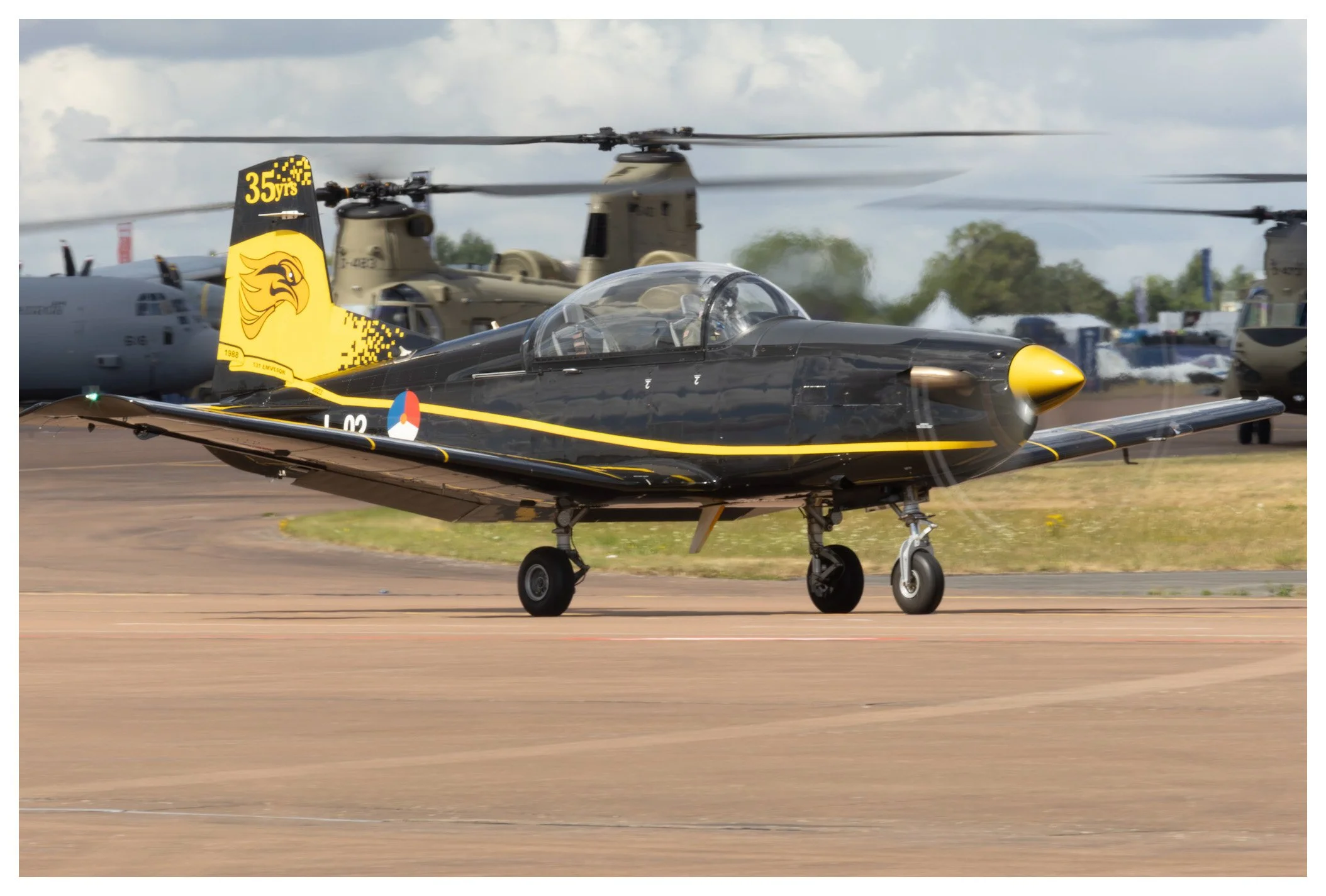 A black aircraft with yellow accents and a yellow tail fin featuring a lion logo, taxis on a runway with helicopters and vehicles in the background.