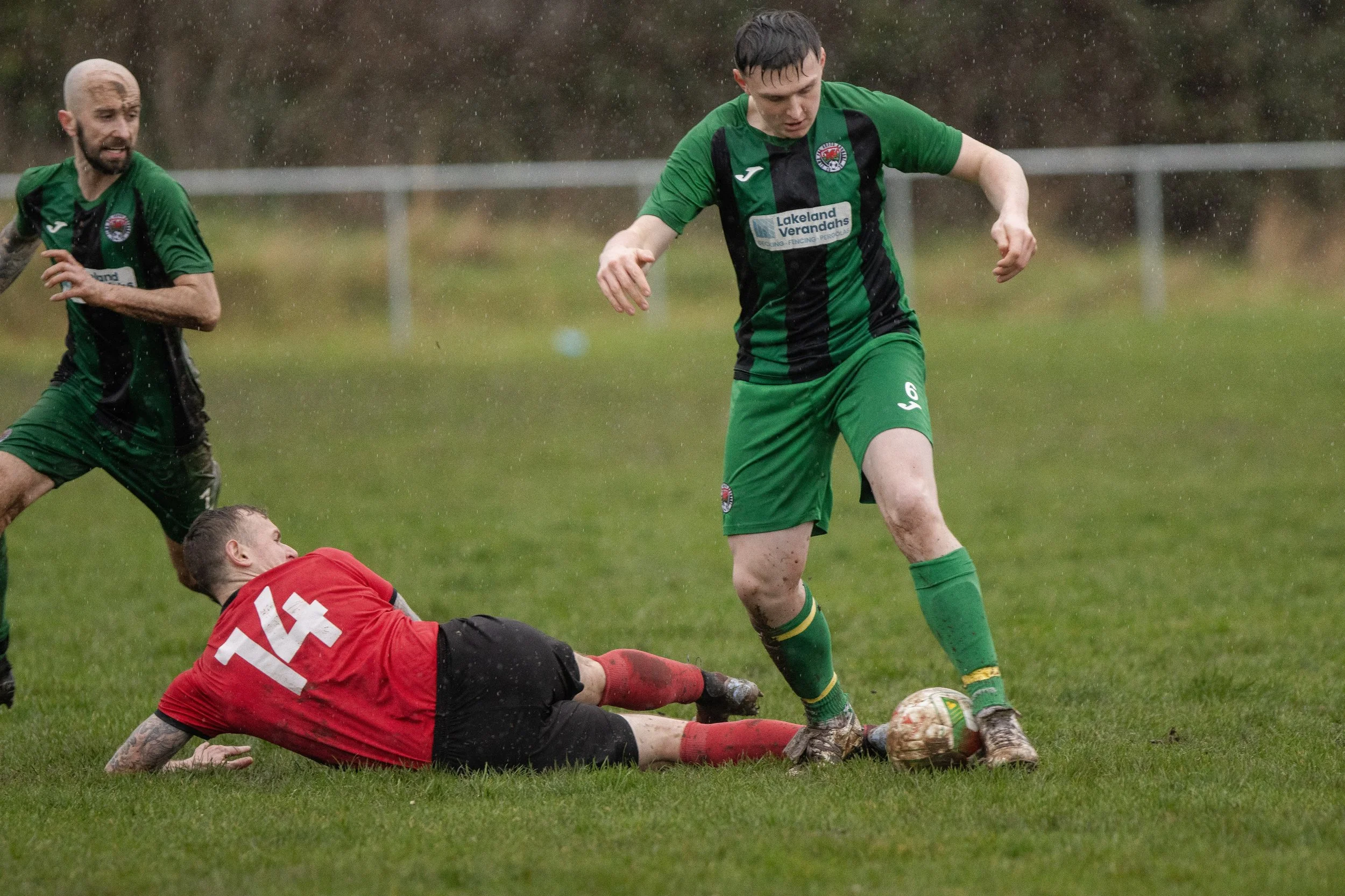 Three soccer players on a field during a rainy game; one player in a red jersey is on the ground, two players in green jerseys are standing, with one of them controlling the ball.