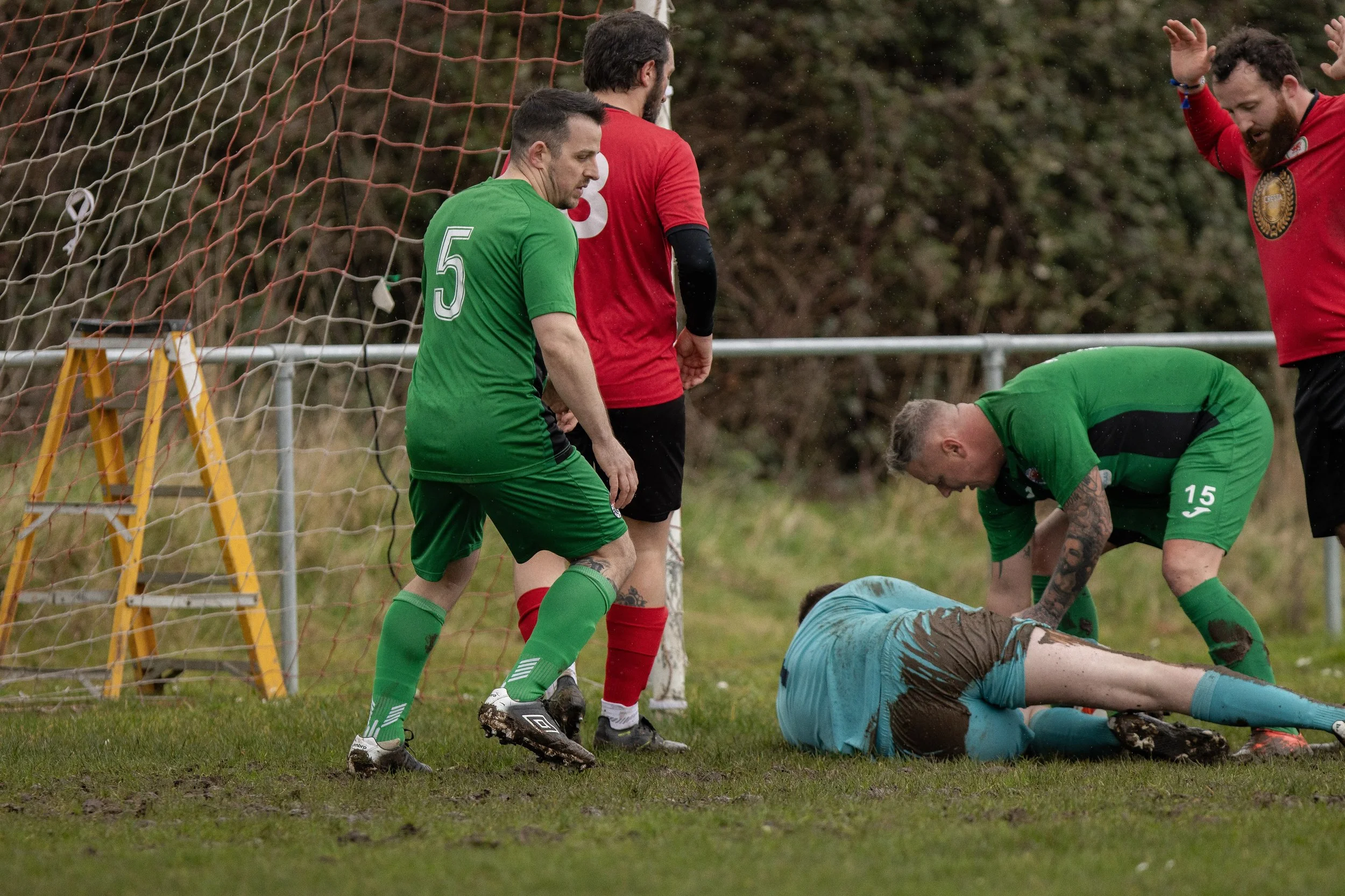 Soccer players in green and red uniforms assist an injured player on the ground near the goal, with one player bending over and another standing nearby.
