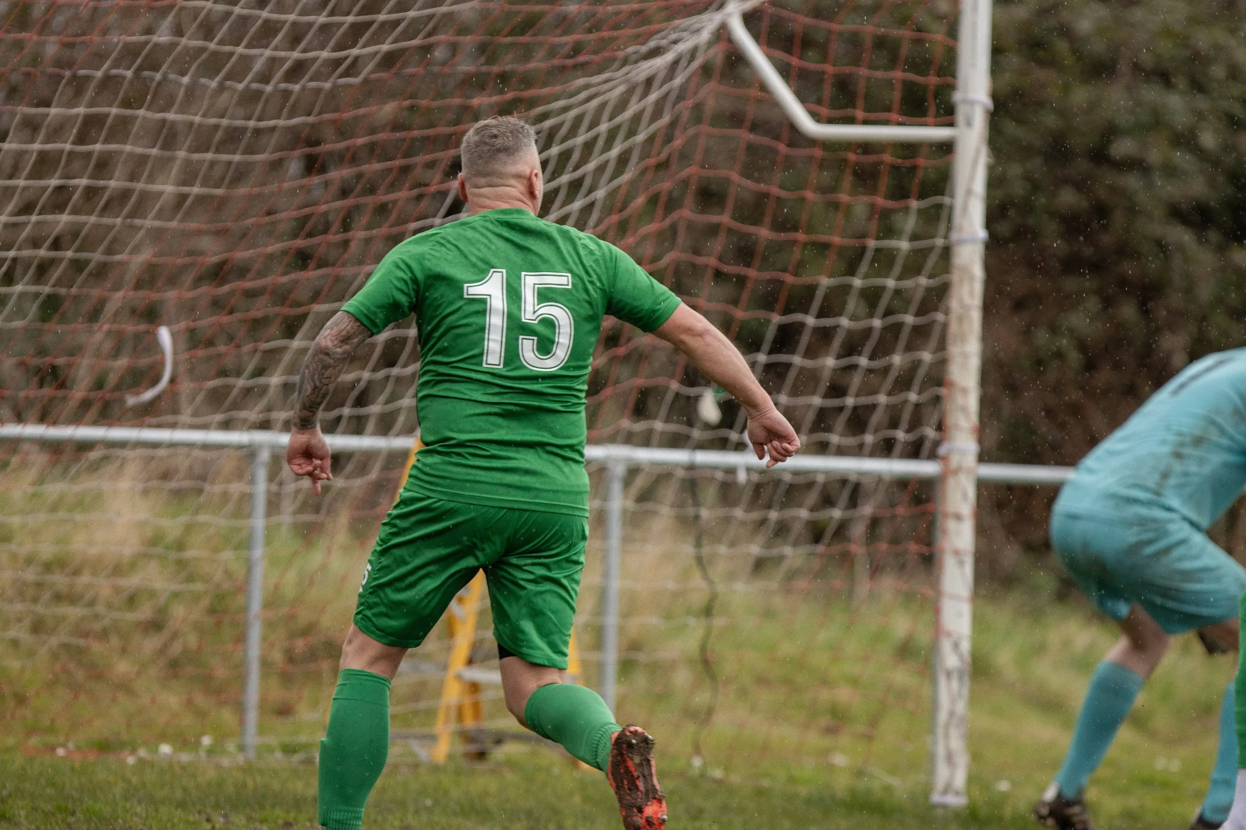A soccer player wearing a green jersey with the number 15 kicks the ball in front of a goal, with an opposing goalie in teal in the background.