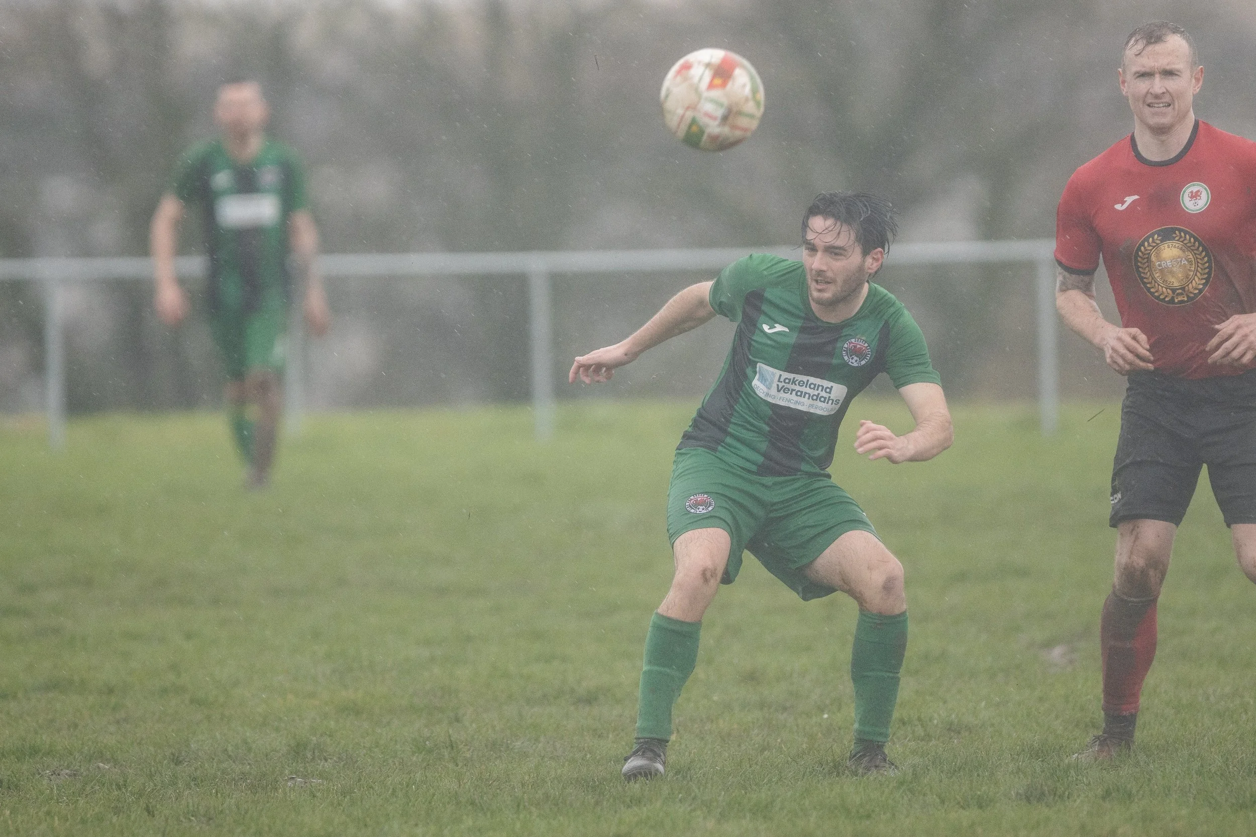 A soccer match in heavy rain with players in green and red uniforms. A player in green is heading the ball, with another player in red nearby, on a wet grassy field.