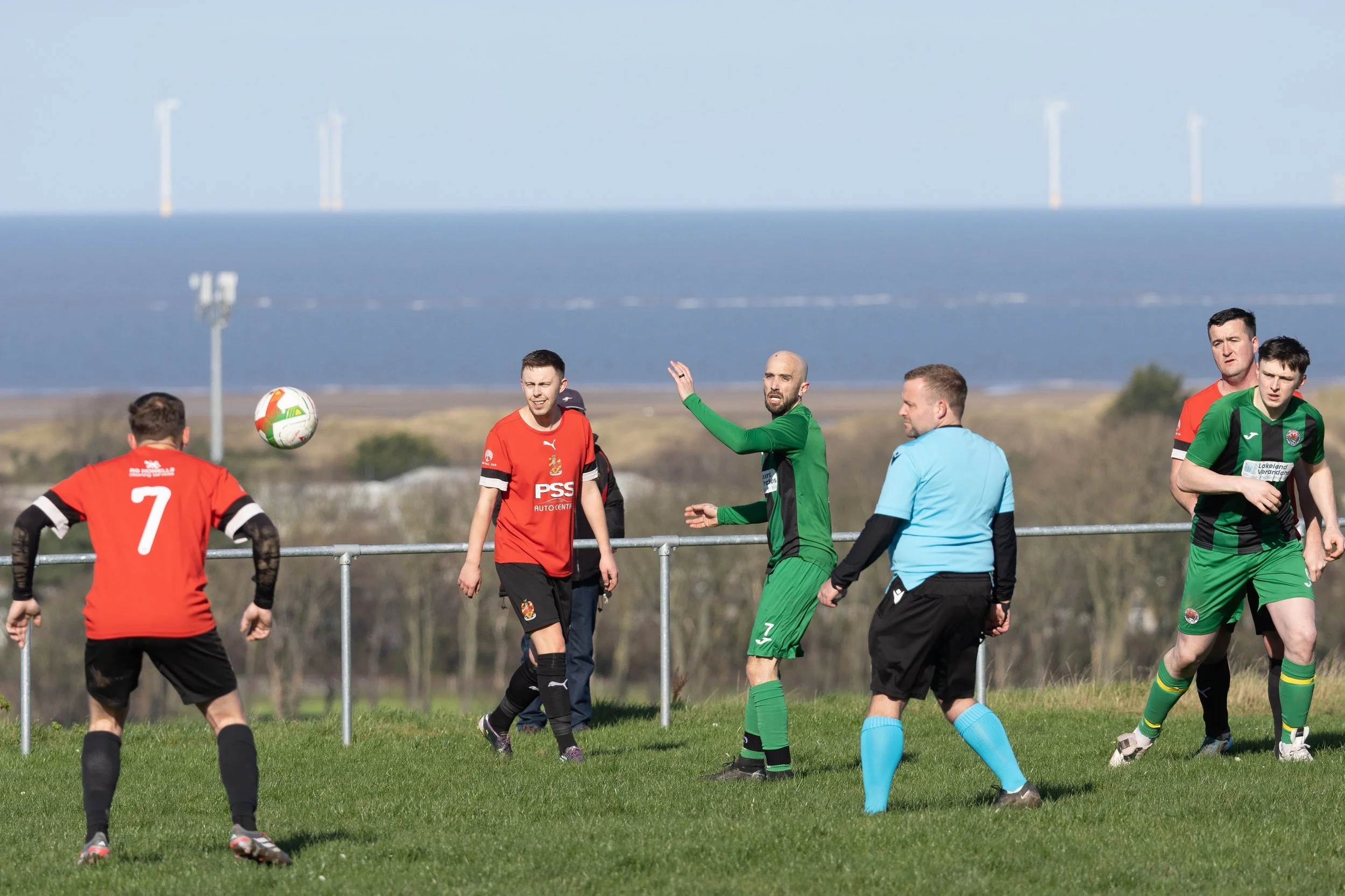 Soccer players in red and green jerseys playing a match on a grassy field with water and wind turbines in the background.