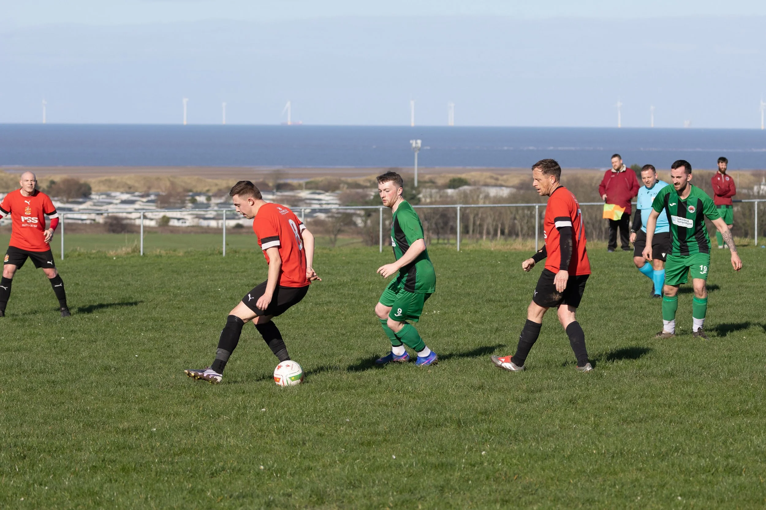Soccer match on a grassy field with players in red and green uniforms, with a coastal landscape and wind turbines in the background.
