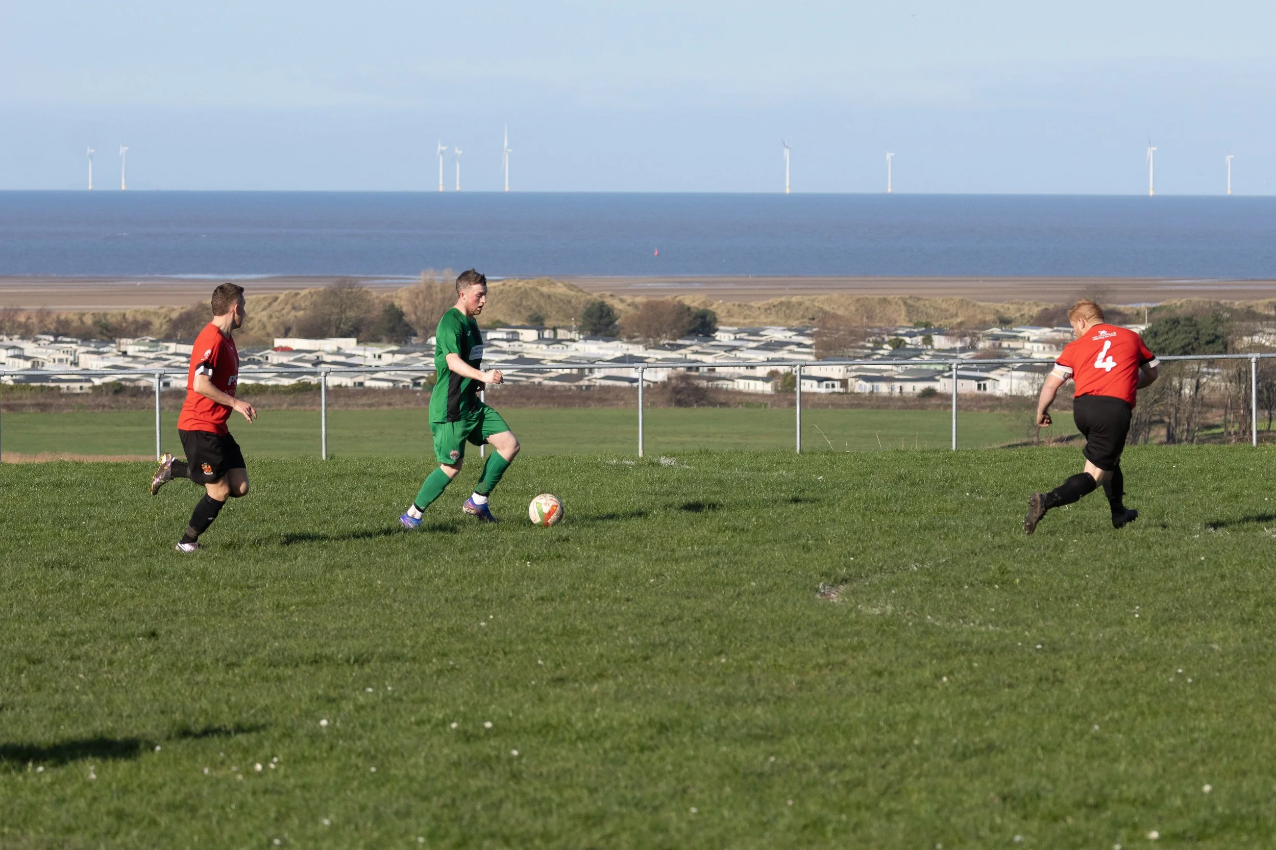 Soccer game with three players on the field near the coast, two in red jerseys and one in a green jersey, with wind turbines in the background.