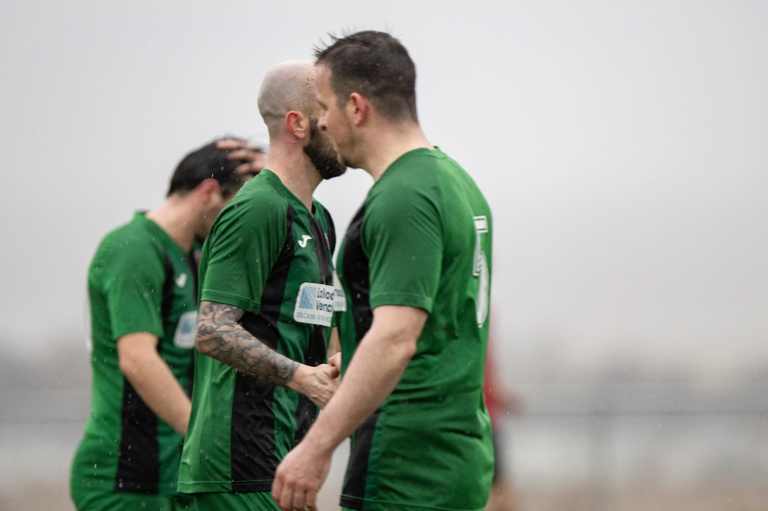 Two soccer players in green jerseys face each other on a rainy day, touching foreheads during a moment of celebration or encouragement, with two other players in the background.