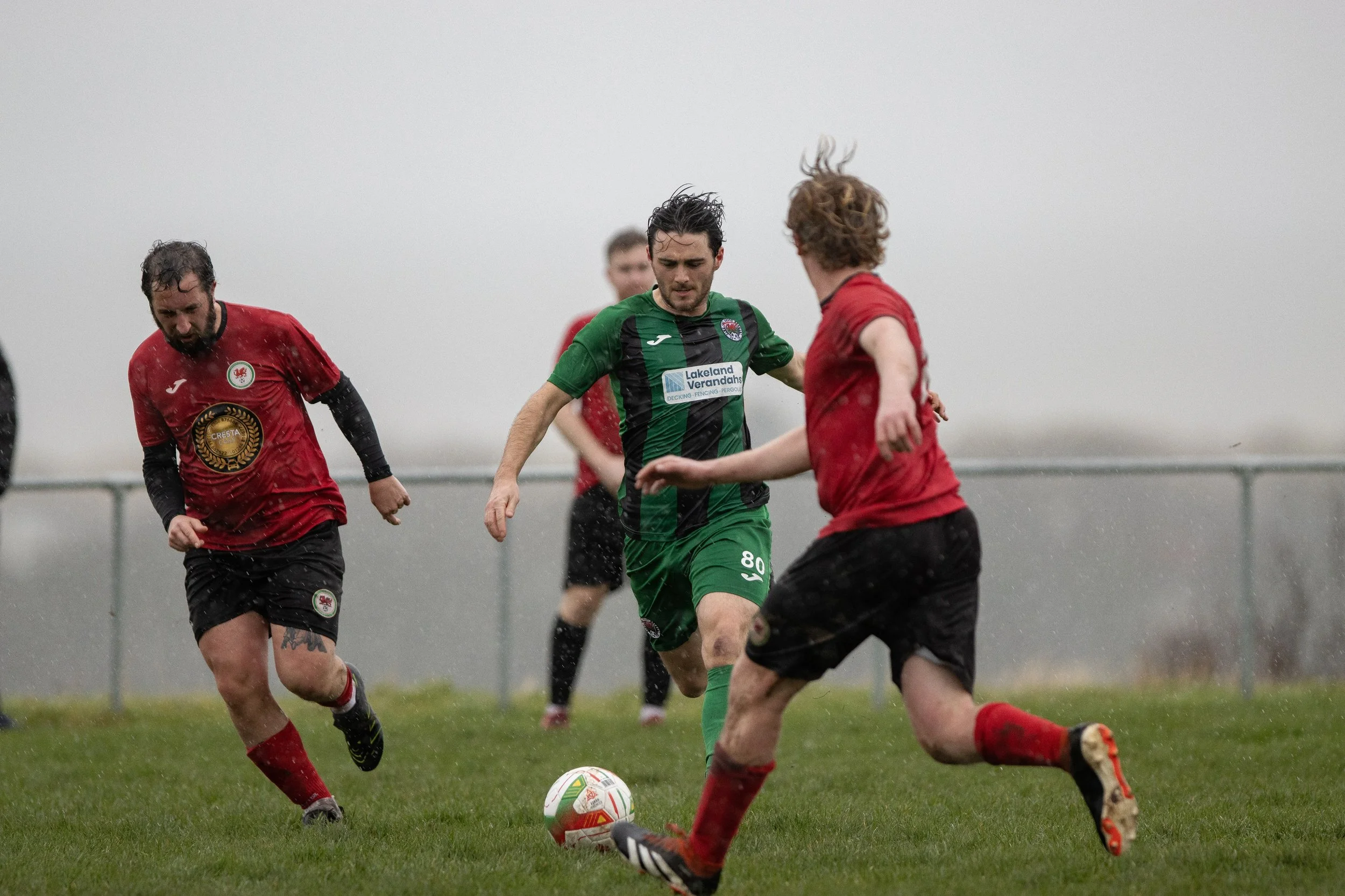 Soccer players on a rainy field competing for the ball, wearing red and green jerseys.