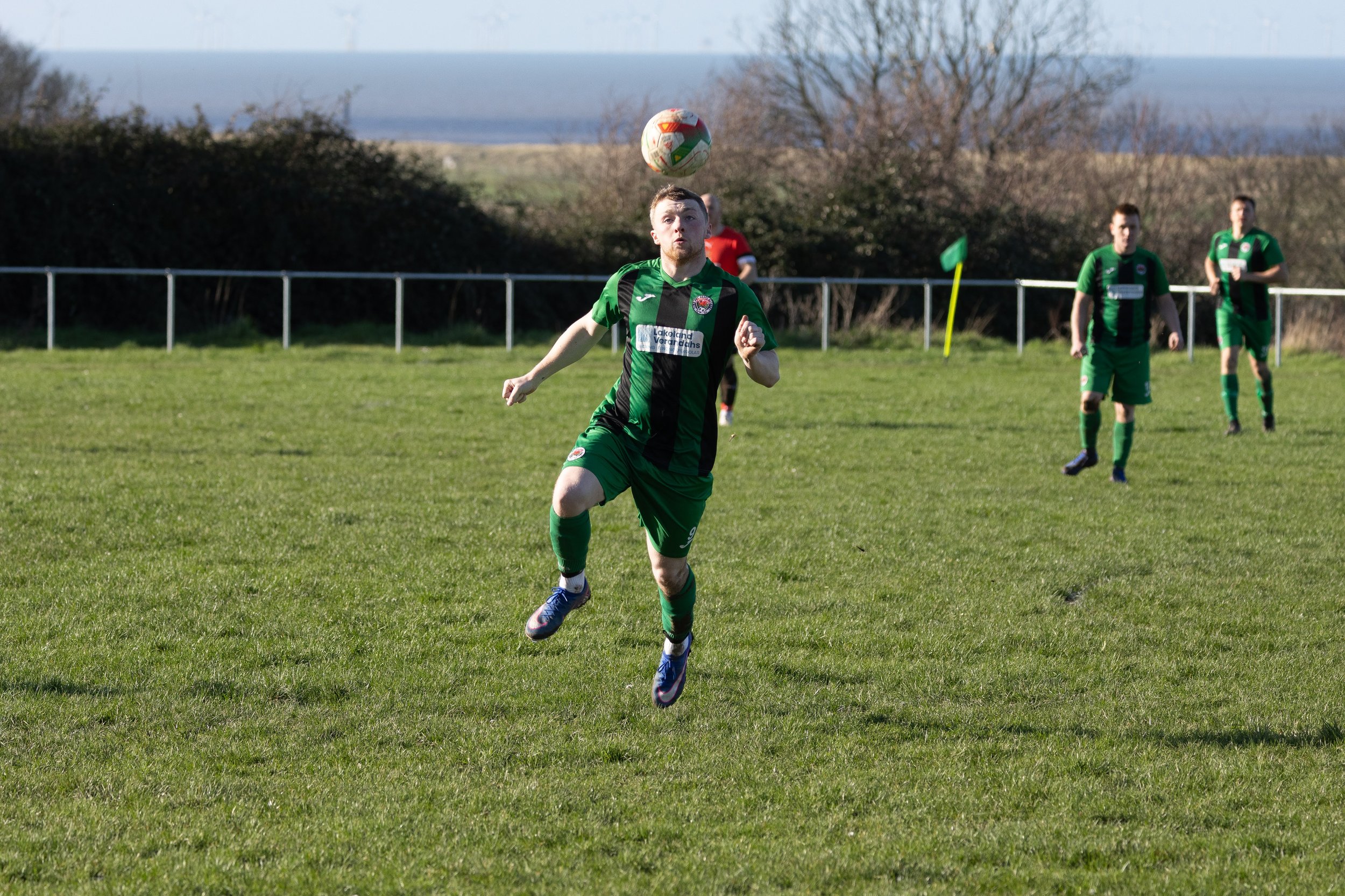 A soccer player in a green and black uniform runs on the field as a soccer ball approaches overhead, with other players and a corner flag visible in the background.