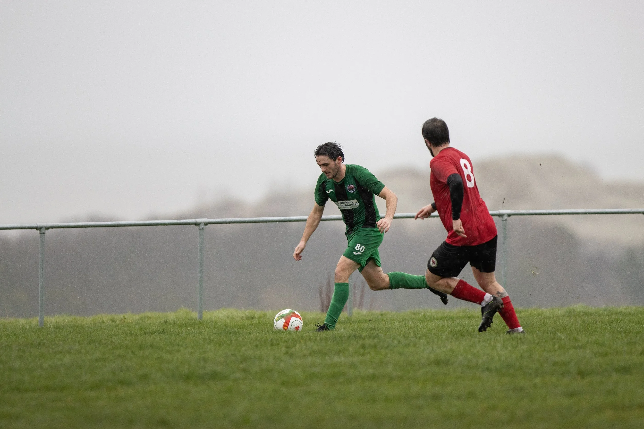 Two soccer players competing for the ball on a grassy field in rainy weather, with a metal railing in the background.