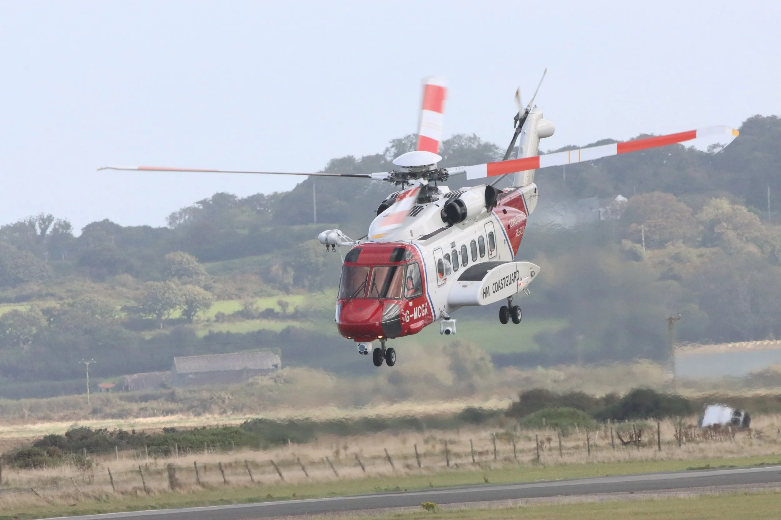 A helicopter hovering above the ground near a rural area with grass, trees, and small buildings in the background.