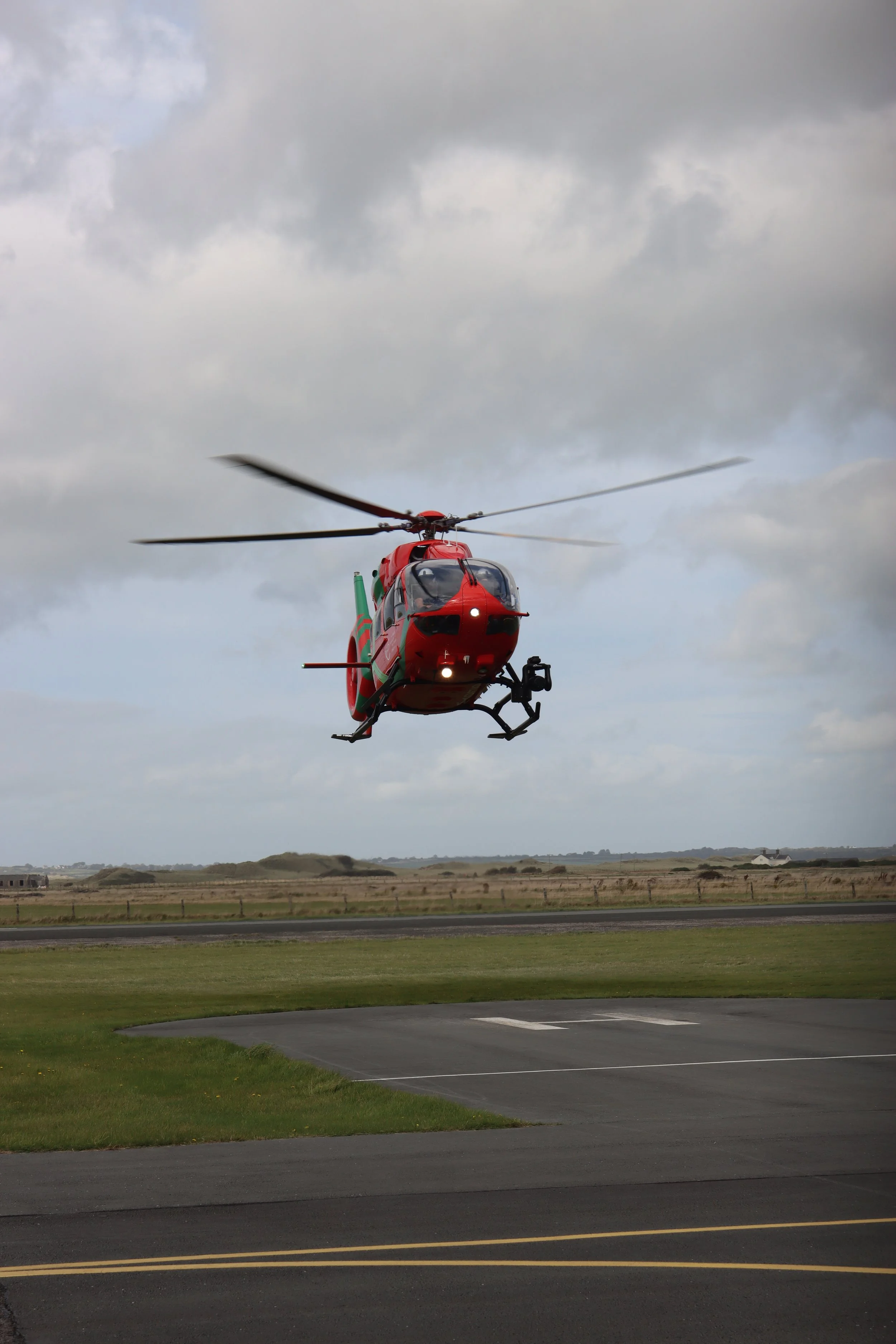 A red helicopter in midair over an open field with cloudy sky in the background.