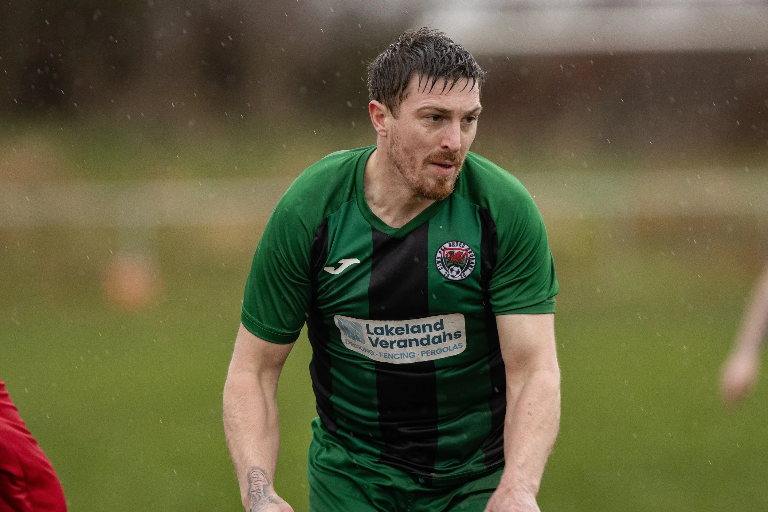 A male soccer player in a green and black jersey with the logo of Club Dreu Drefu on the chest, playing outdoors on a grassy field in rainy weather.
