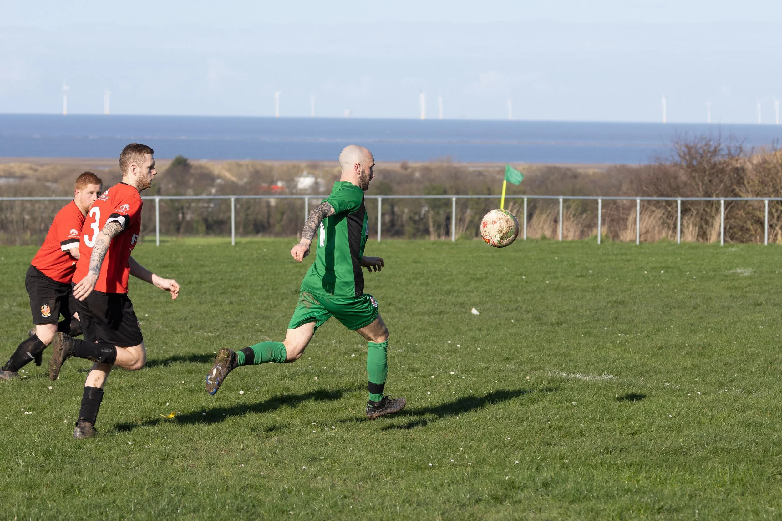 Soccer players chasing the ball on a grassy field with a fence, trees, and wind turbines in the background.