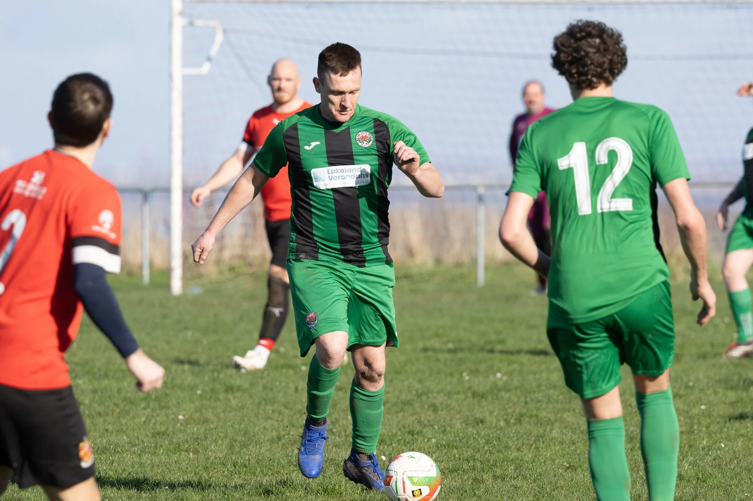 Soccer players on the field during a match, with one player in a green and black uniform controlling the ball while others in green and red uniforms watch.