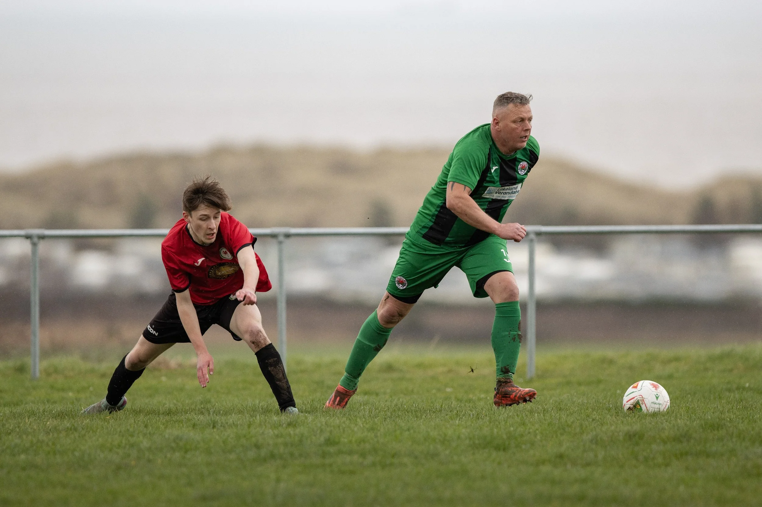 Two soccer players, one in a red jersey and the other in a green jersey, competing for the ball on a grassy field with cloudy skies.