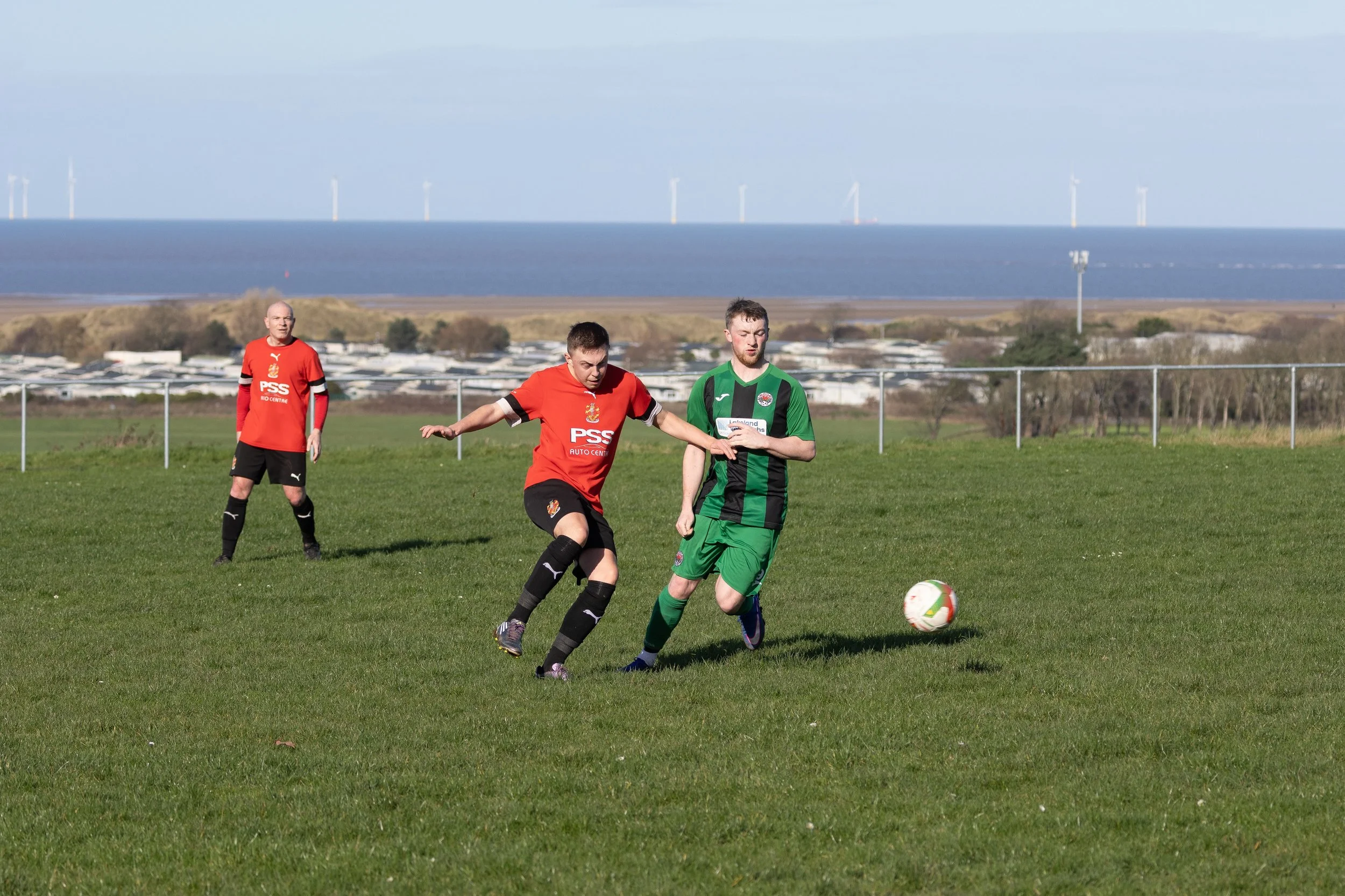 Three soccer players on a grassy field with the ocean and wind turbines in the background. Two players in red jerseys and one in a green jersey are competing for the soccer ball.