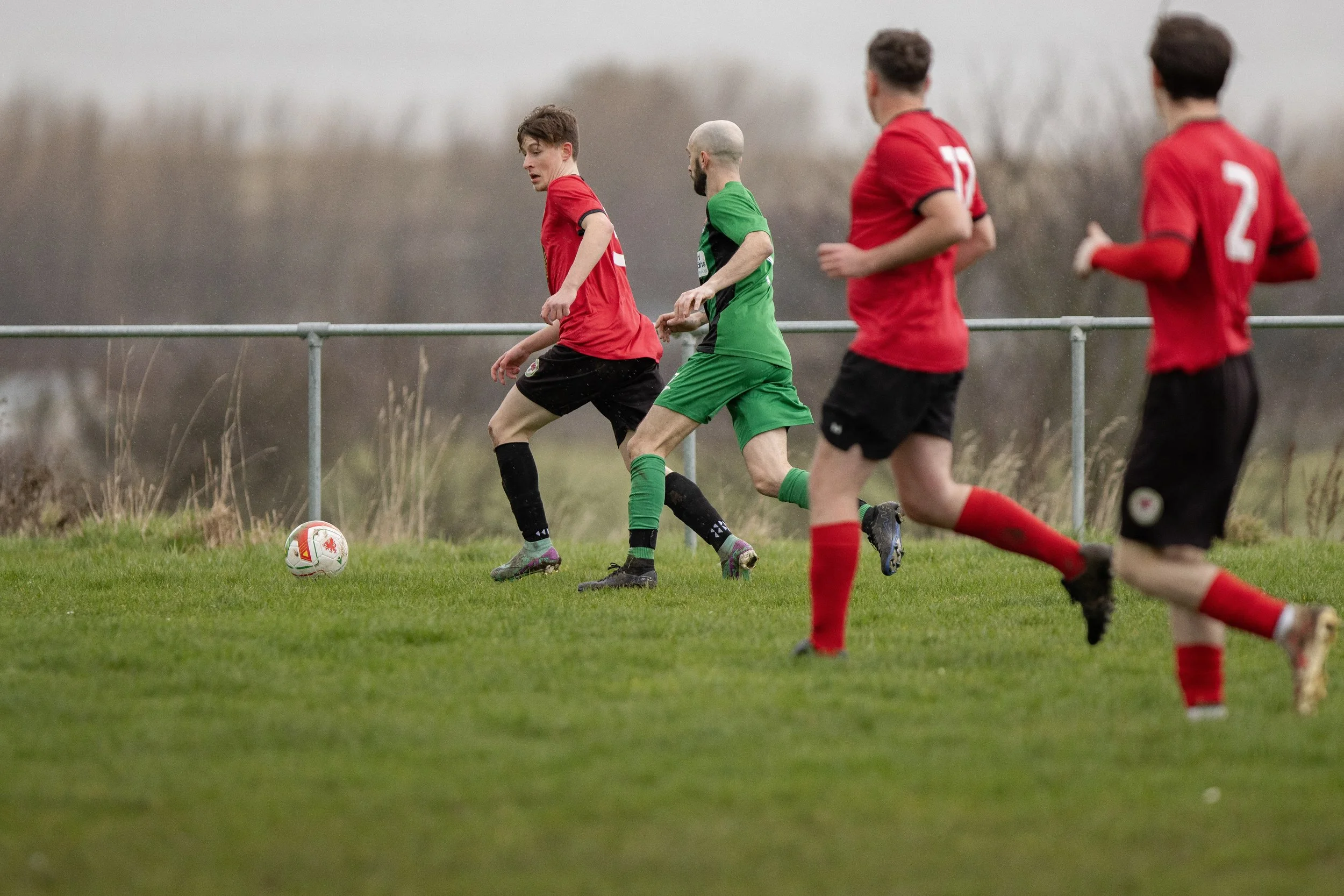 Soccer players in red and green uniforms playing on a grass field, with one player controlling the ball as others run nearby.