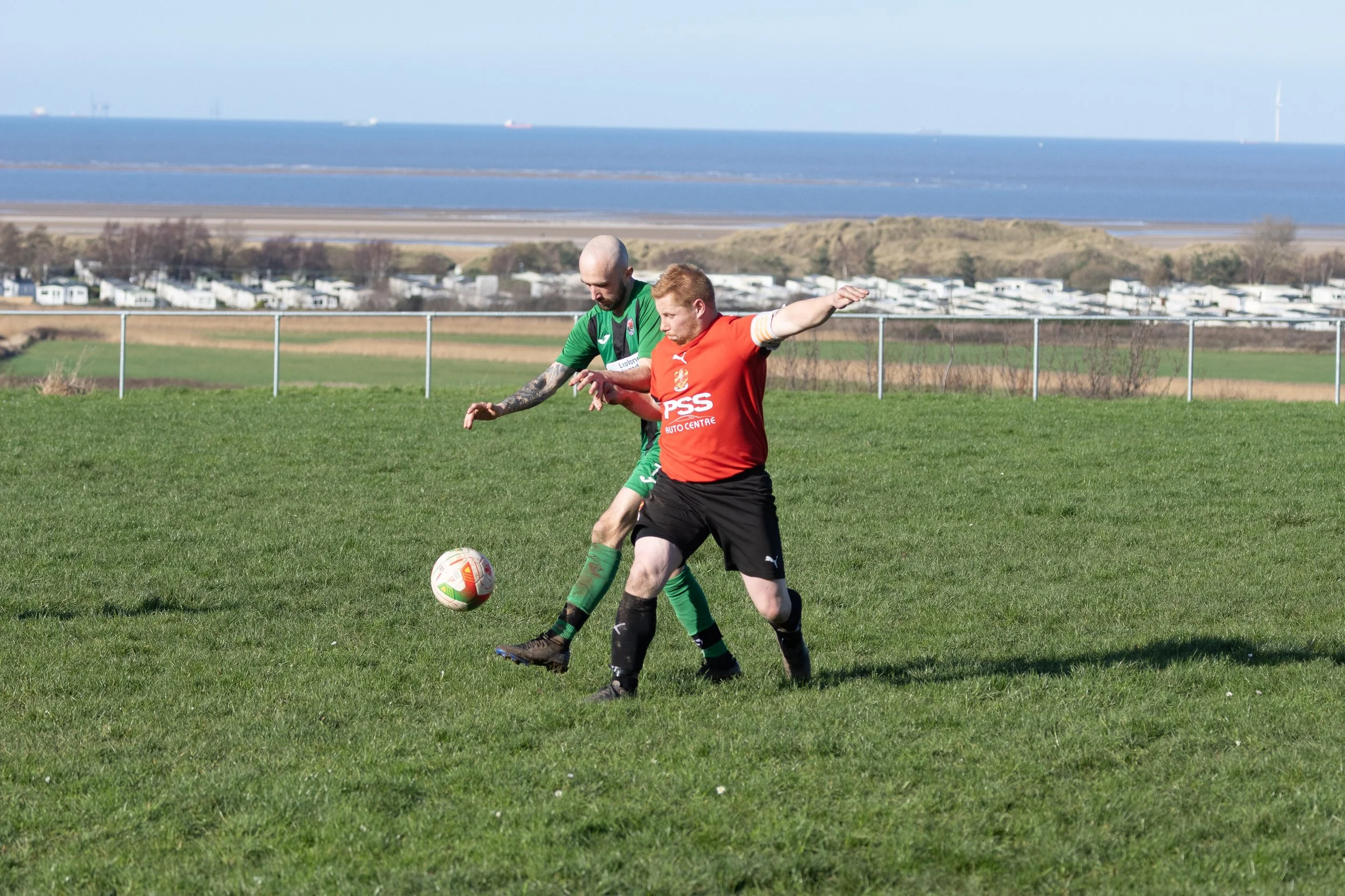Two soccer players competing for ball on grassy field, with ocean and distant landscape in background.
