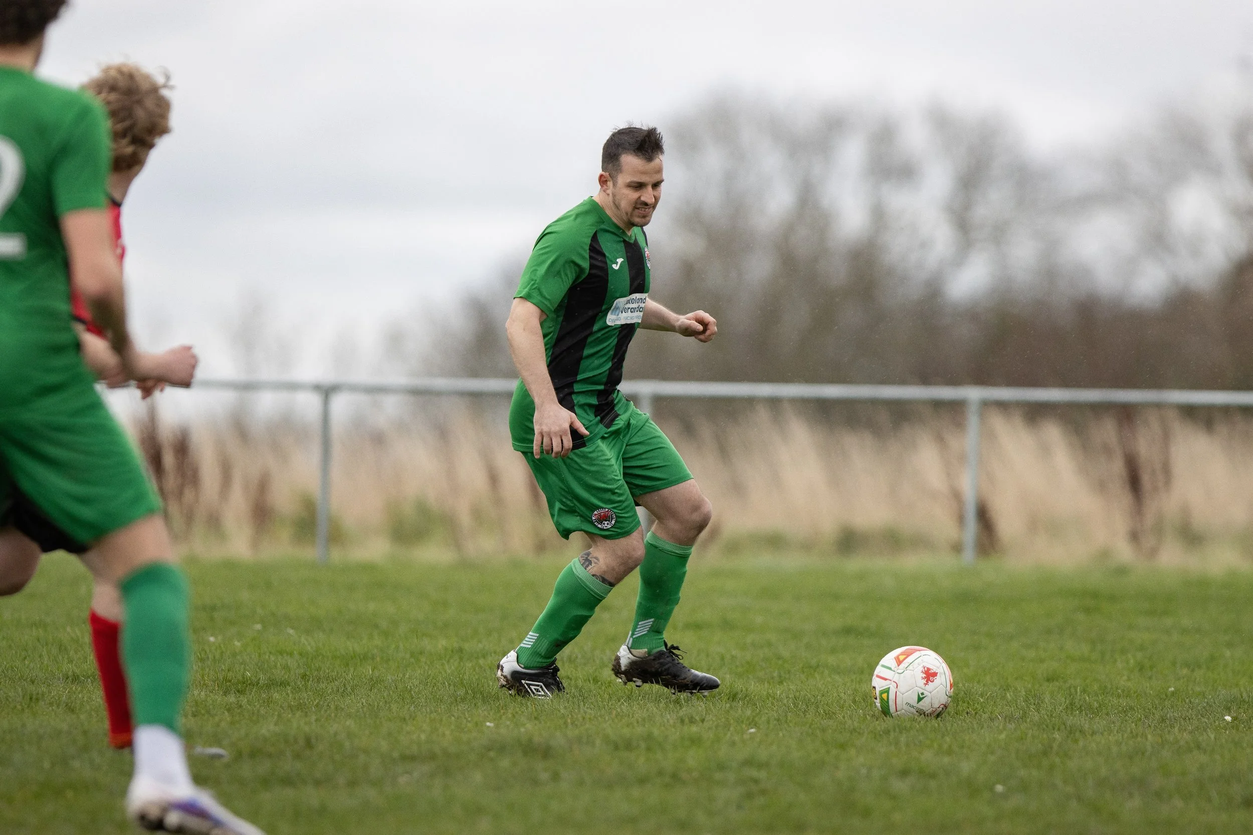 A man in a green and black soccer jersey and shorts, with green socks and black cleats, playing soccer on a grassy field during an overcast day, with other players partially visible in the background.