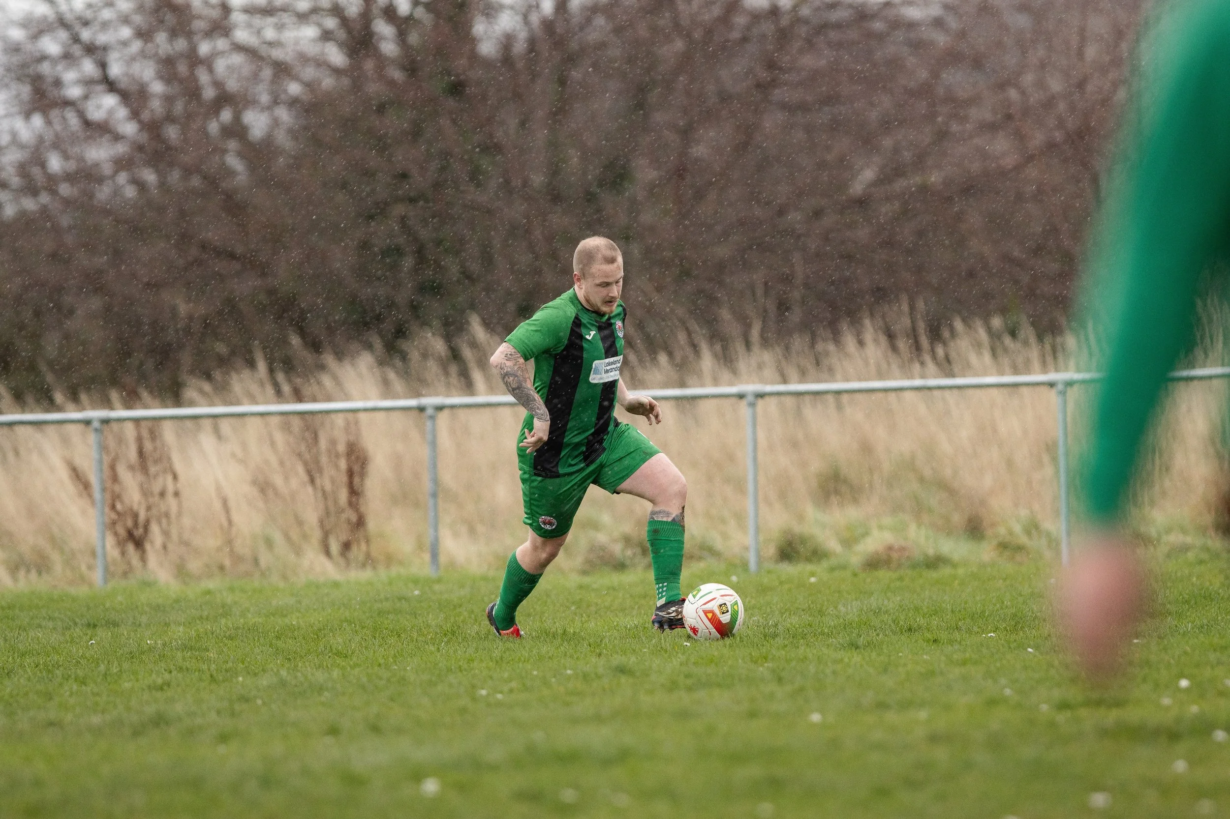 A soccer player in a green and black uniform prepares to kick a soccer ball on a grassy field, with a metal fence and trees with brown leaves in the background.
