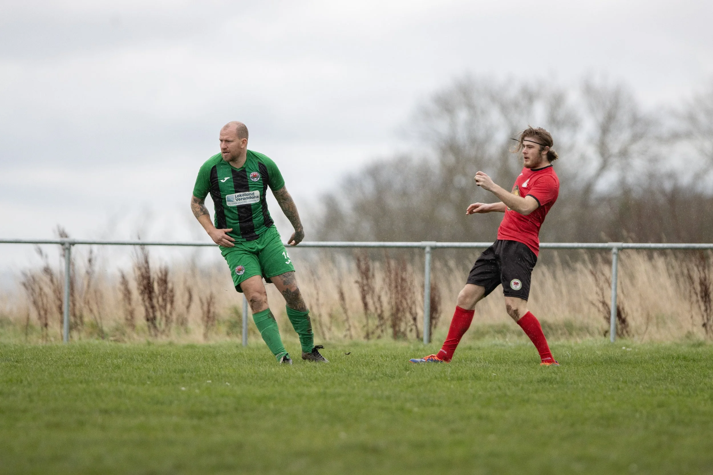 Two male soccer players on a grass field, one in a green and black uniform running and the other in a red and black uniform preparing for a kick, with a chain-link fence and trees in the background.
